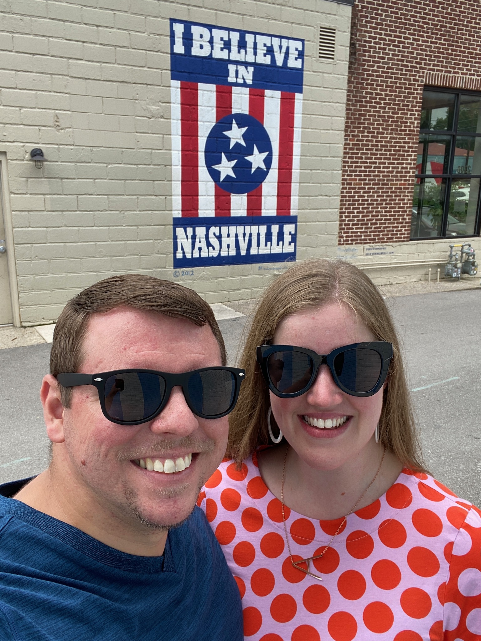 Two people wearing sunglasses pose in front of a mural that reads "I BELIEVE IN NASHVILLE" with stars and stripes in the background.