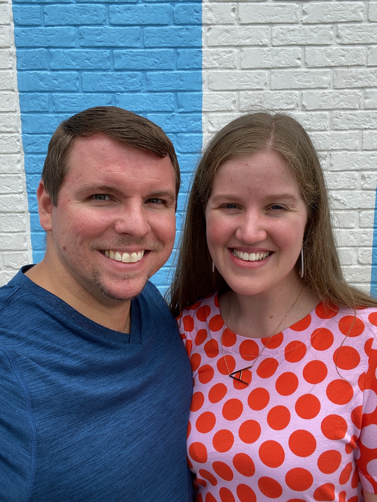 A man in a blue shirt and a woman in a red polka-dot shirt smile in front of a blue and white brick wall.