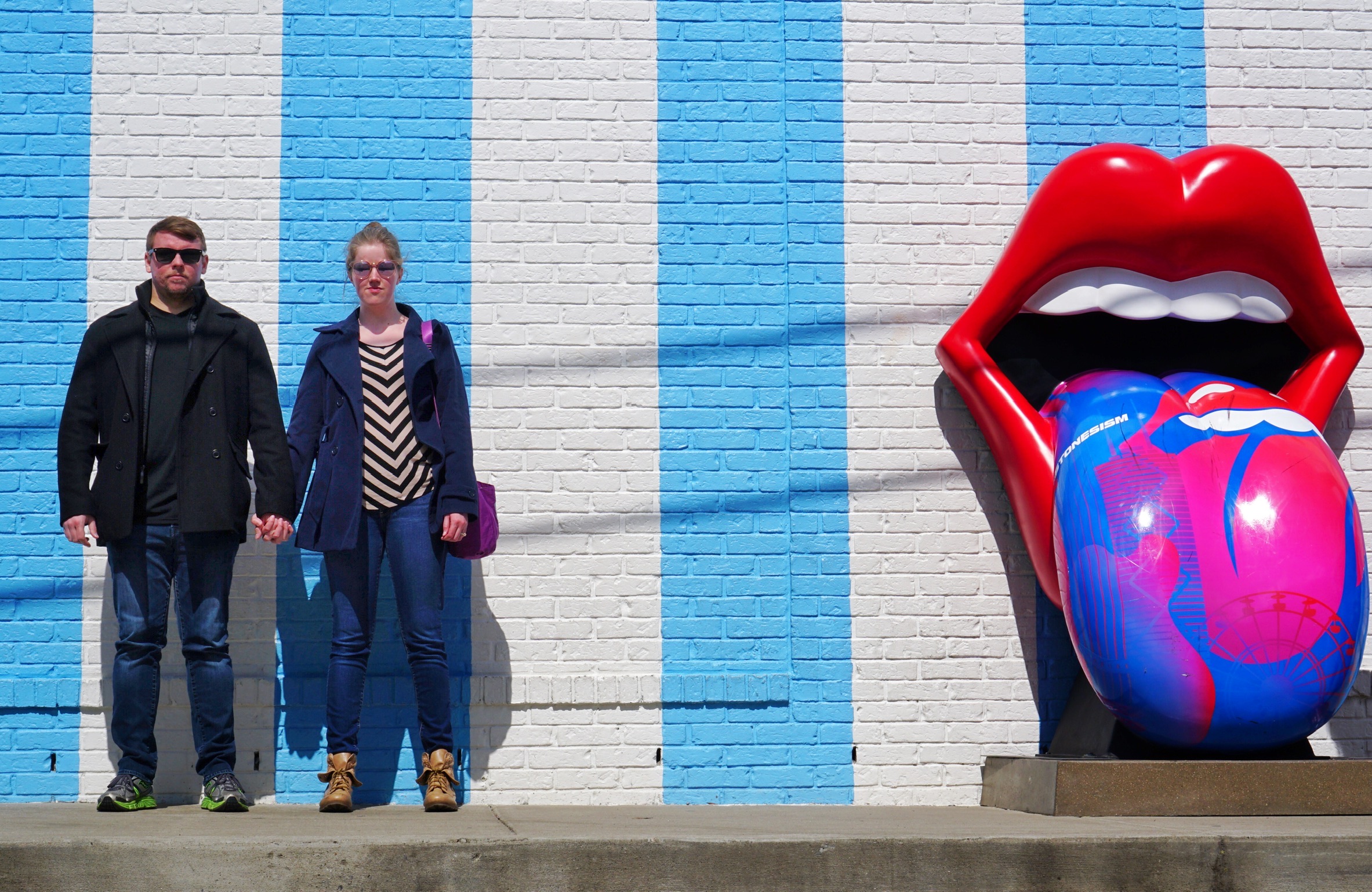 Two people stand holding hands in front of a white brick wall with blue stripes, next to a large sculpture of red lips with a colorful tongue.