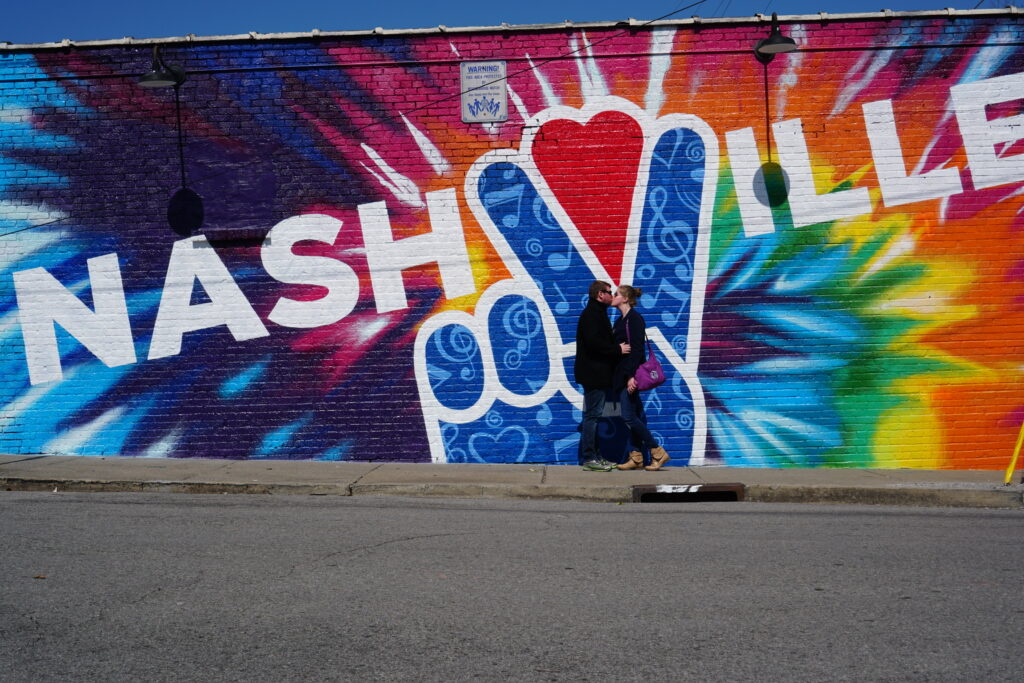 Two people stand in front of a colorful Nashville mural featuring a large peace sign and tie-dye background on a brick wall.