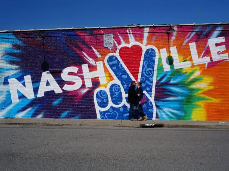 Two people stand in front of a colorful Nashville mural featuring a large peace sign and tie-dye background on a brick wall.