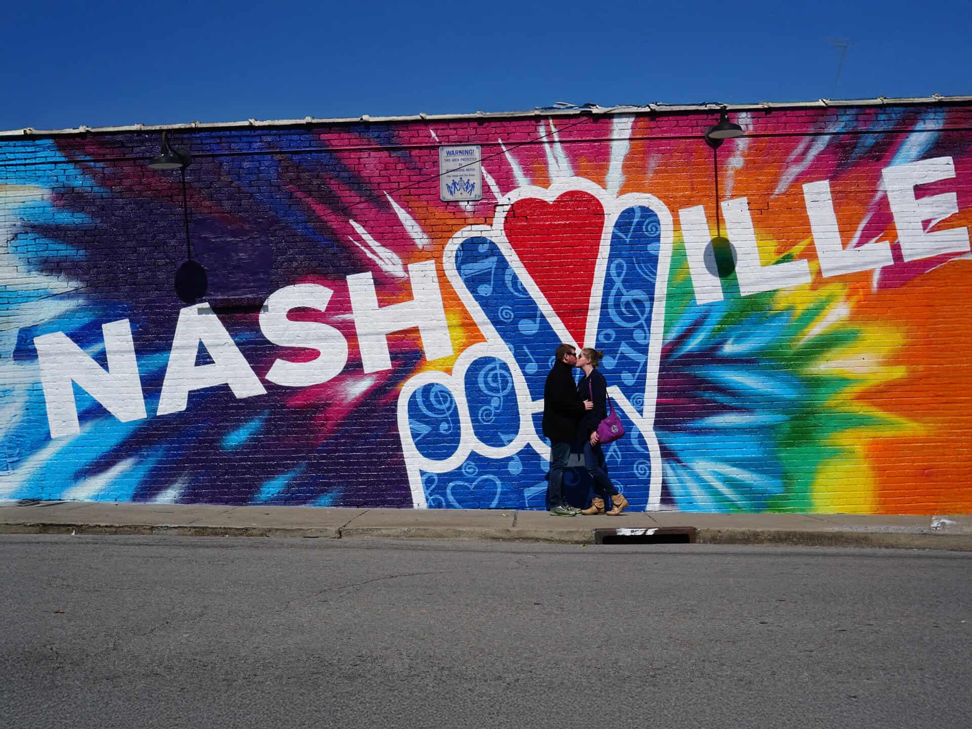Two people stand in front of a colorful Nashville mural featuring a large peace sign and tie-dye background on a brick wall.