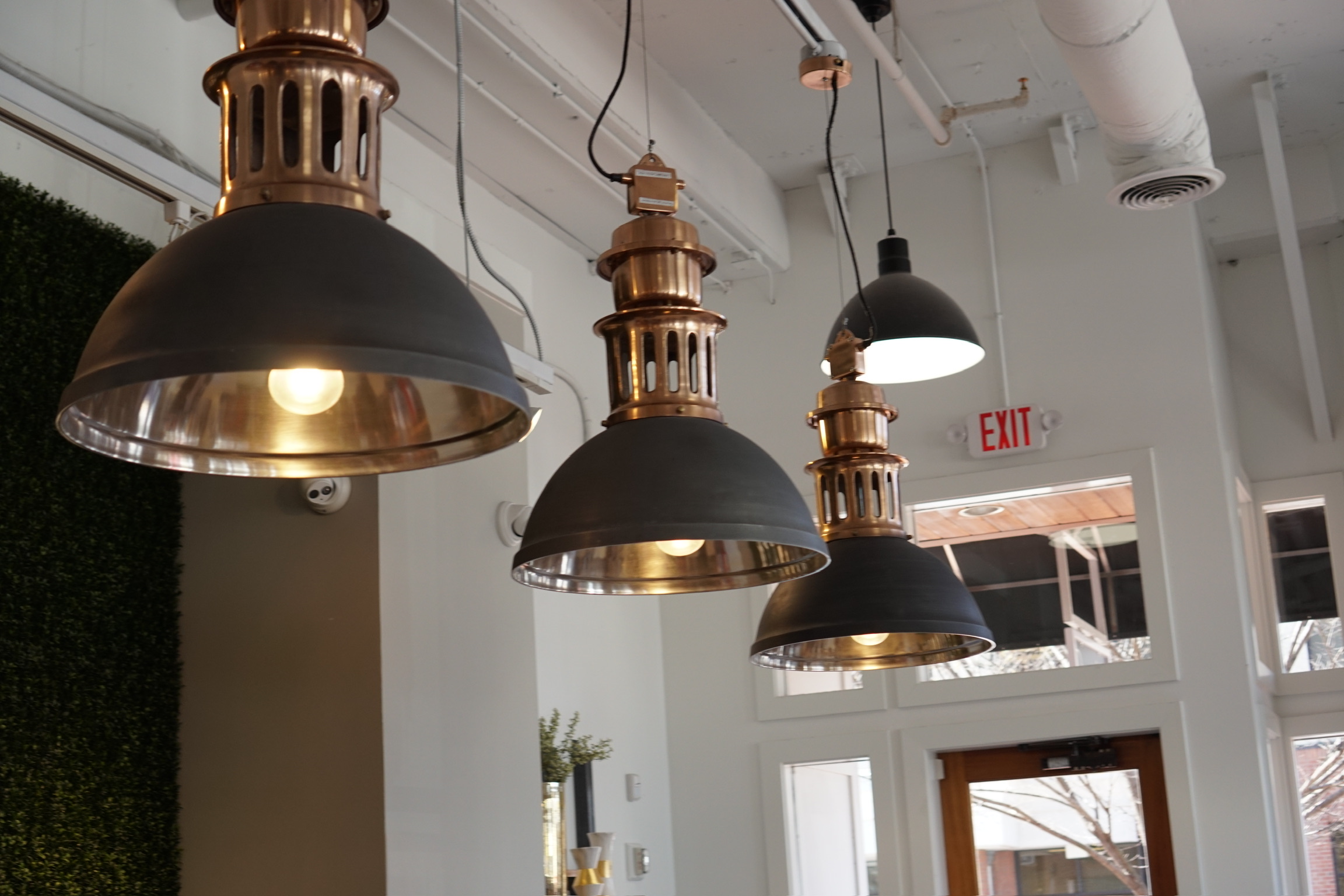 A row of industrial-style pendant lights hangs from the ceiling in a modern interior with large windows and an exit sign.