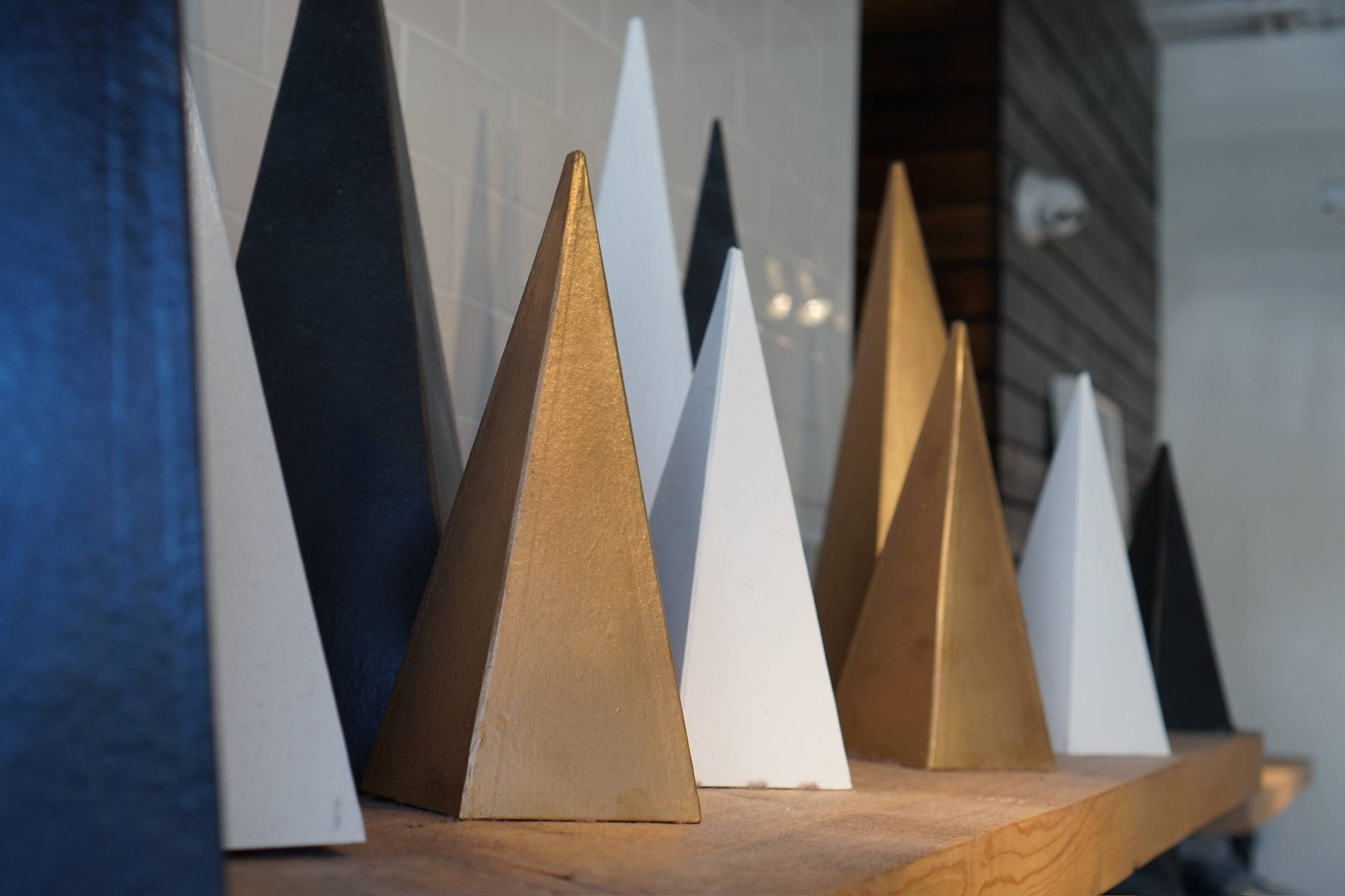 Several pyramid-shaped objects in black, white, and gold are arranged in a row on a wooden shelf against a tiled and wooden wall.