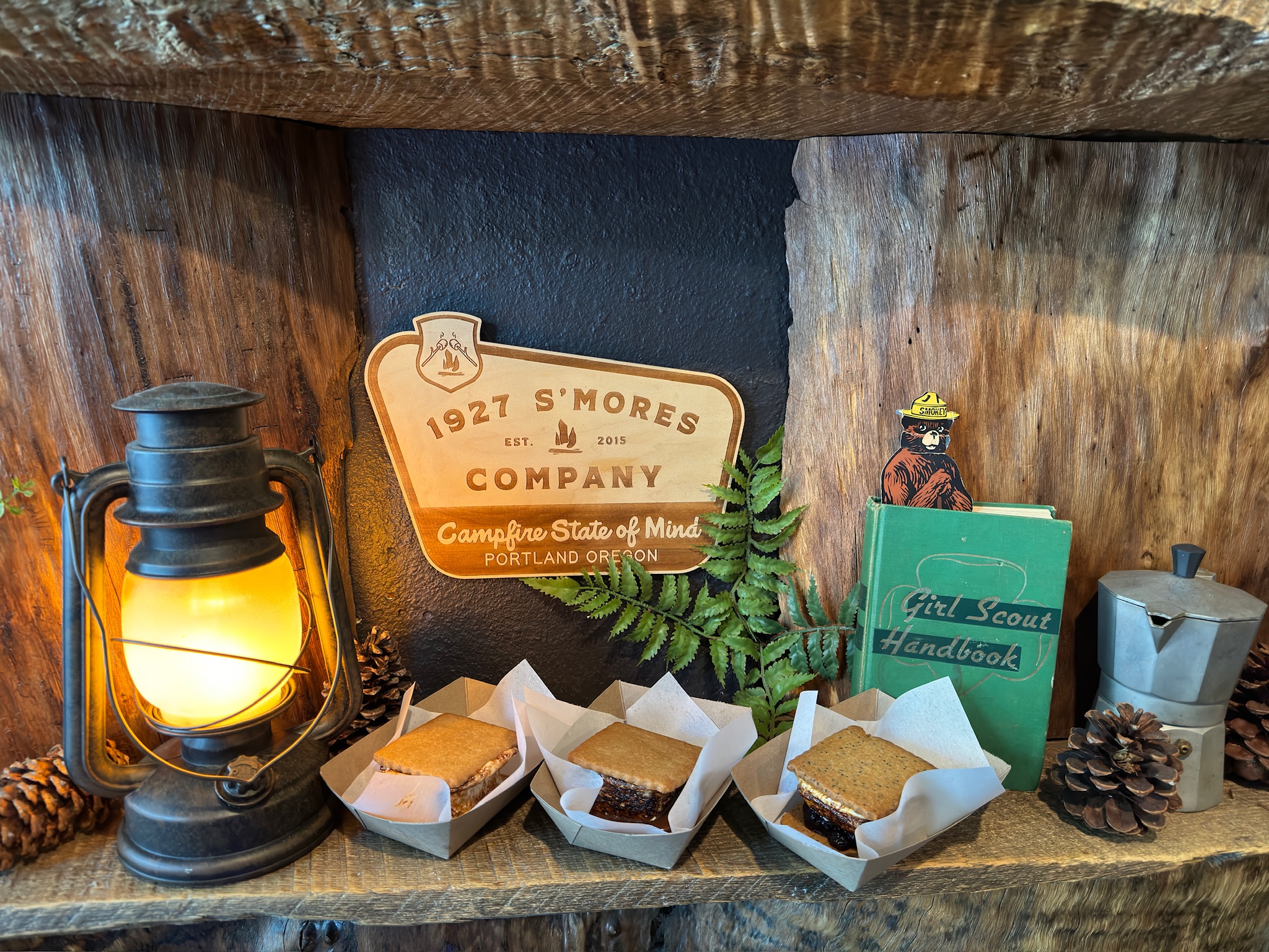 A vintage lantern, three s'mores in paper trays, a "1927 S'mores Company" sign, a Girl Scout Handbook, and pinecones arranged on a rustic wooden shelf.