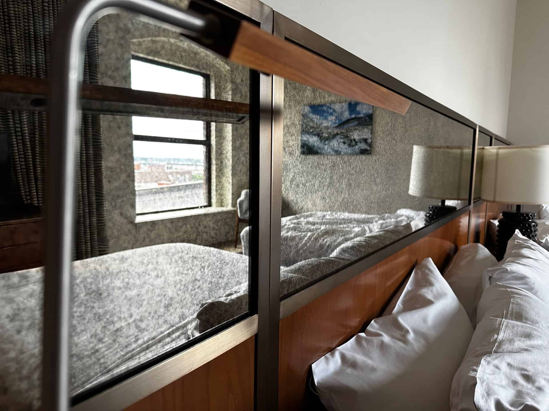 A close-up of a Nashville hotel room's interior featuring a bed with white pillows and a tall headboard with a mirror. A window with curtains and a wall-mounted picture are visible in the reflection.