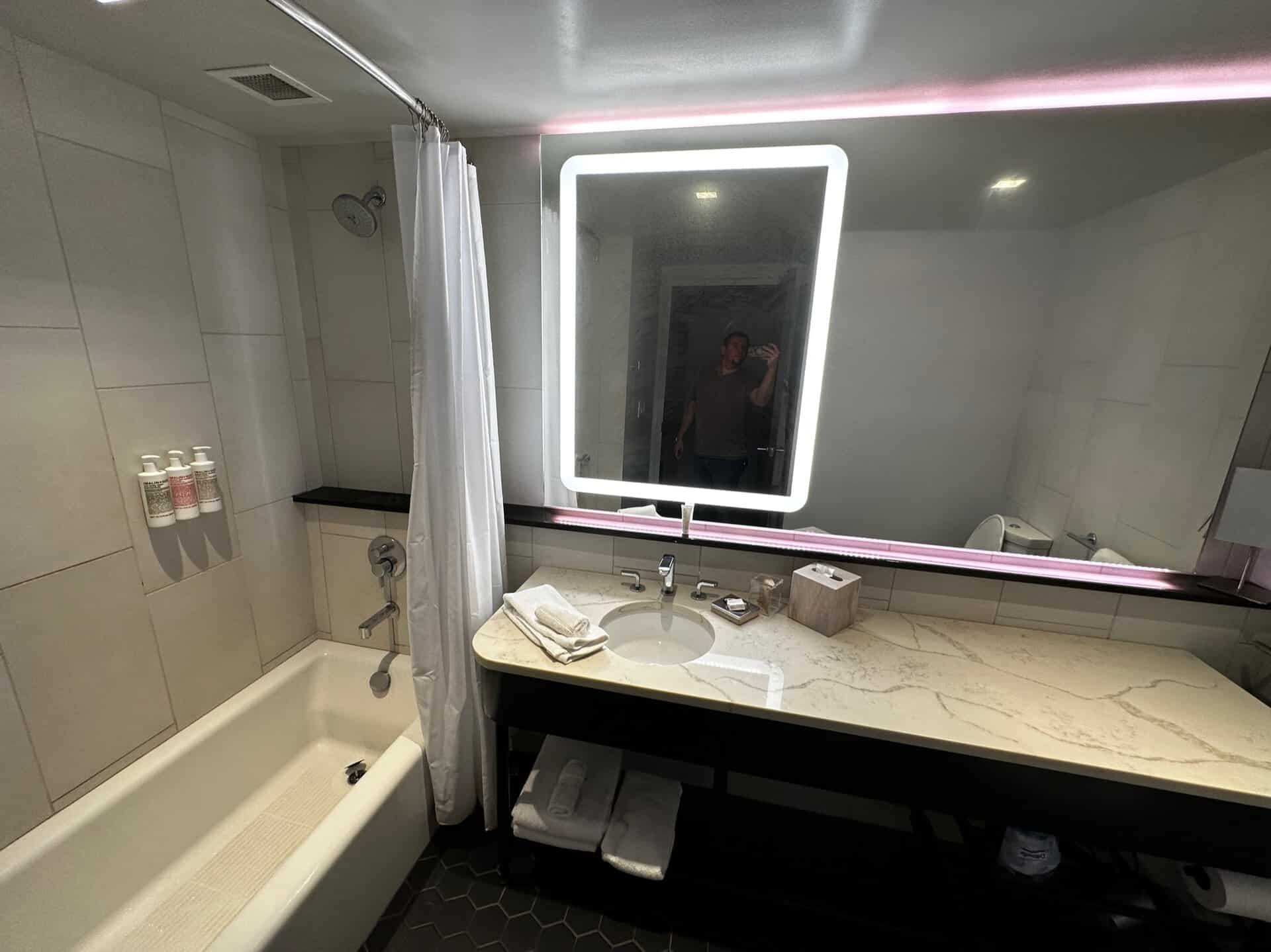 In a Nashville hotel bathroom, a lighted mirror reflects the essentials: sink, toiletries, and a hint of someone adjusting their hat. To the left, the bathtub awaits behind its shower curtain.