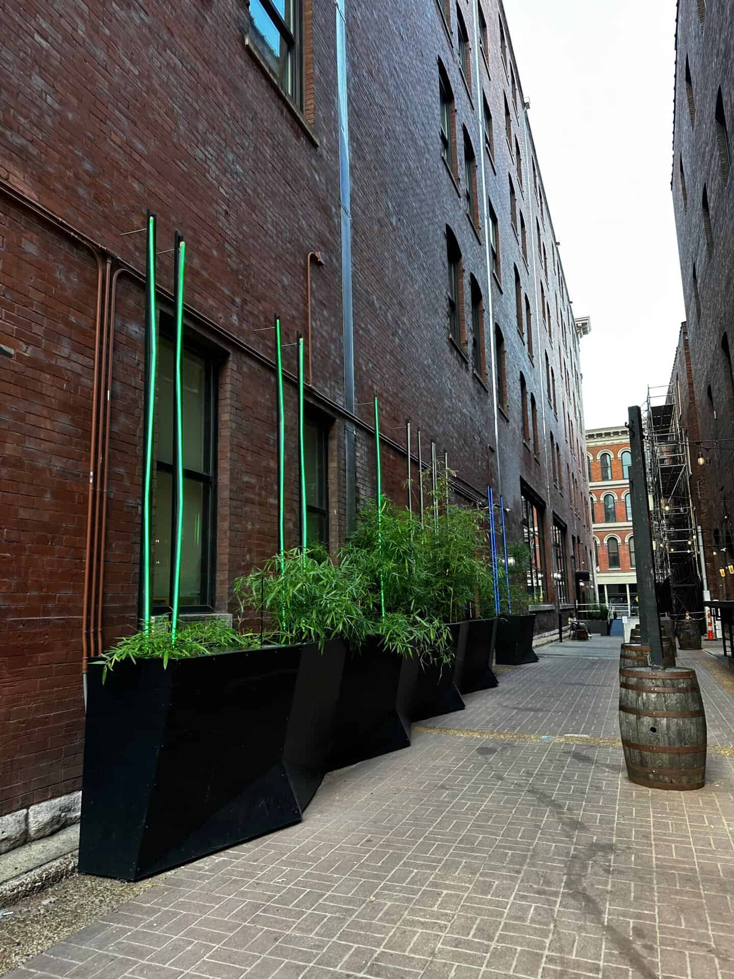 Brick alley with large black planters filled with lush greenery adds a touch of Nashville charm. Colored poles extend from the planters, while a rustic barrel decorates the cobblestone walkway. Adjacent buildings add to the picturesque scene.