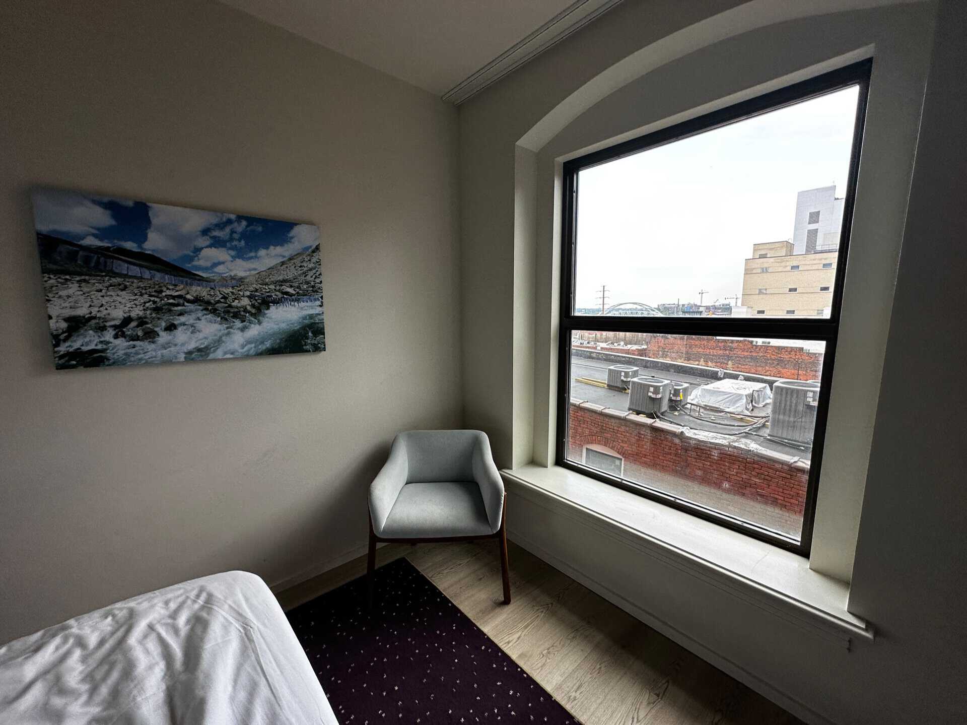 A cozy Nashville bedroom features a bed, an armchair, and a large window that frames the city's skyline. A landscape painting adds charm to the decor, providing a serene contrast to the vibrant urban scene outside.