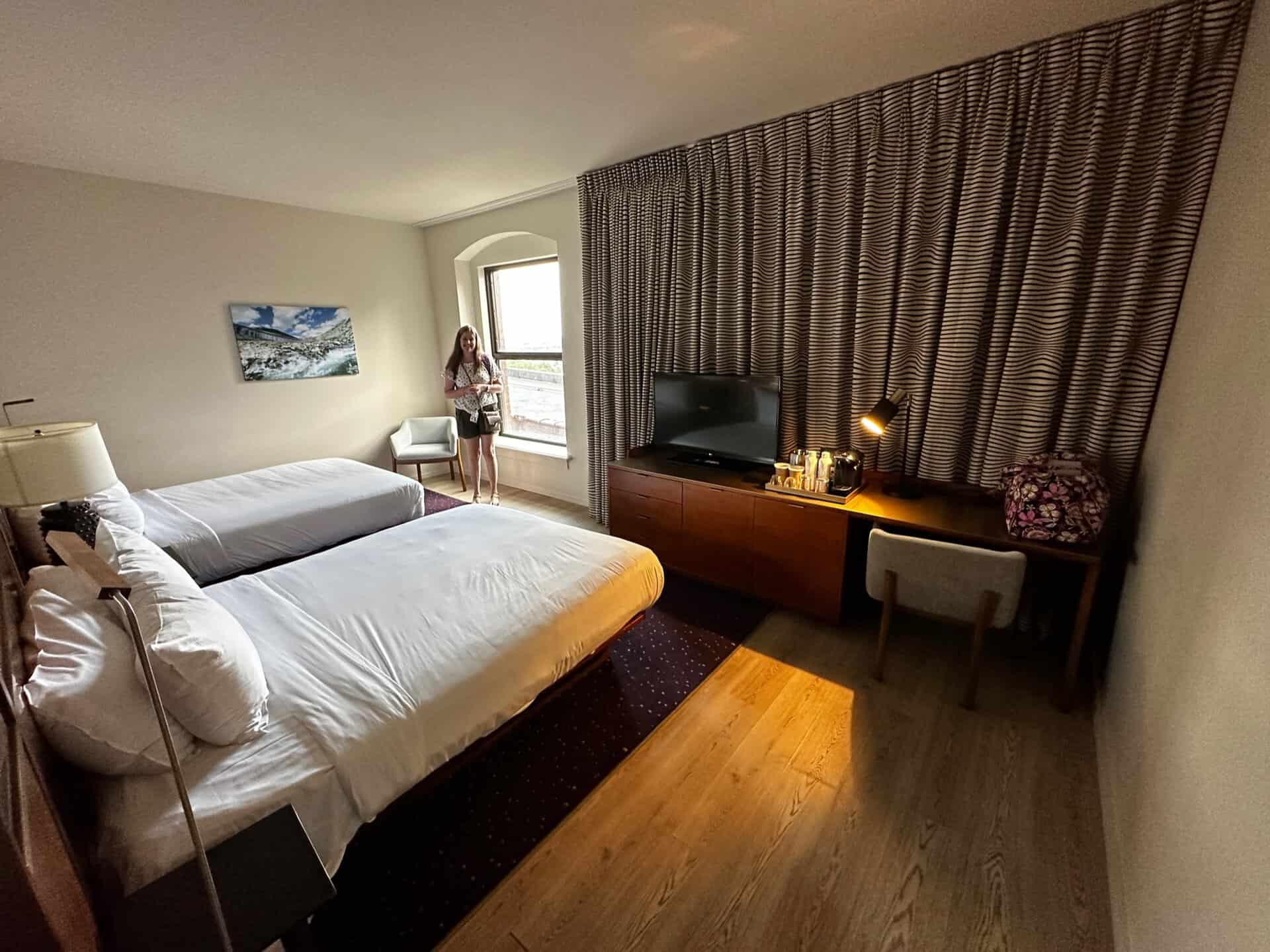 The Nashville hotel room features two beds, a desk with a chair, a TV, and a large window adorned with curtains. A person stands near the window, gazing at the vibrant cityscape.