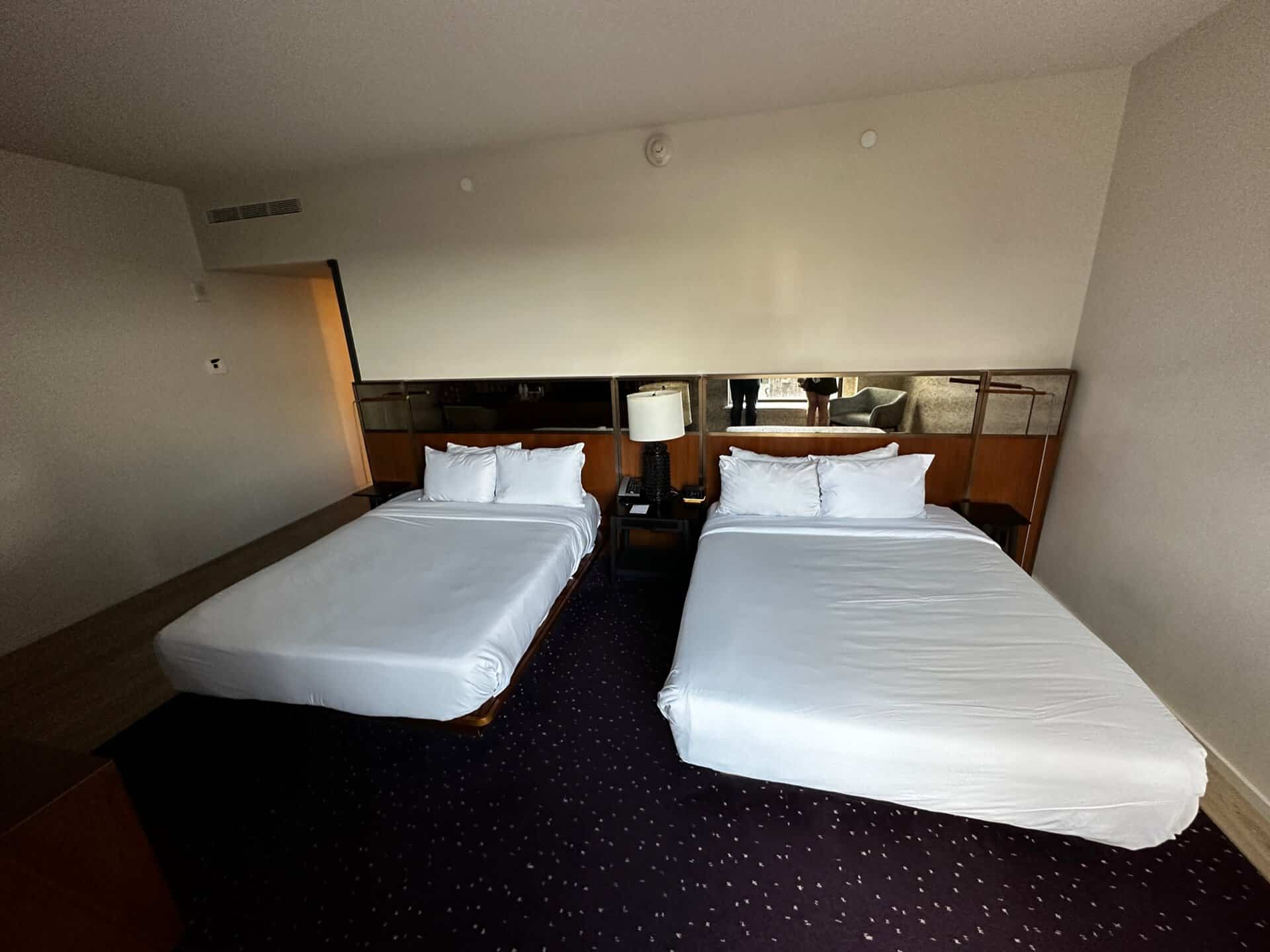 This Nashville hotel room features two neatly made double beds, a cozy bedside table with a lamp between them, and a dark carpeted floor.