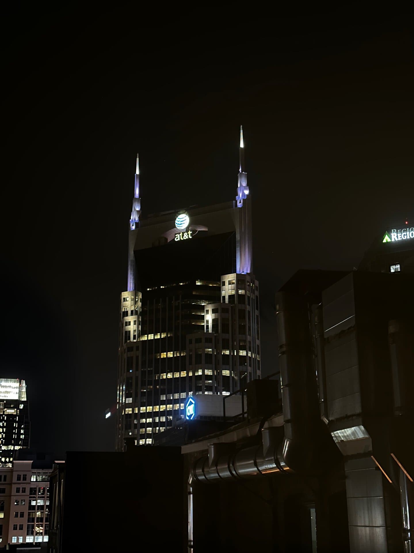 The night view of Nashville's AT&T Building dazzles, showcasing its iconic twin spires brilliantly illuminated against the dark sky, surrounded by glimpses of the city's architectural marvels.