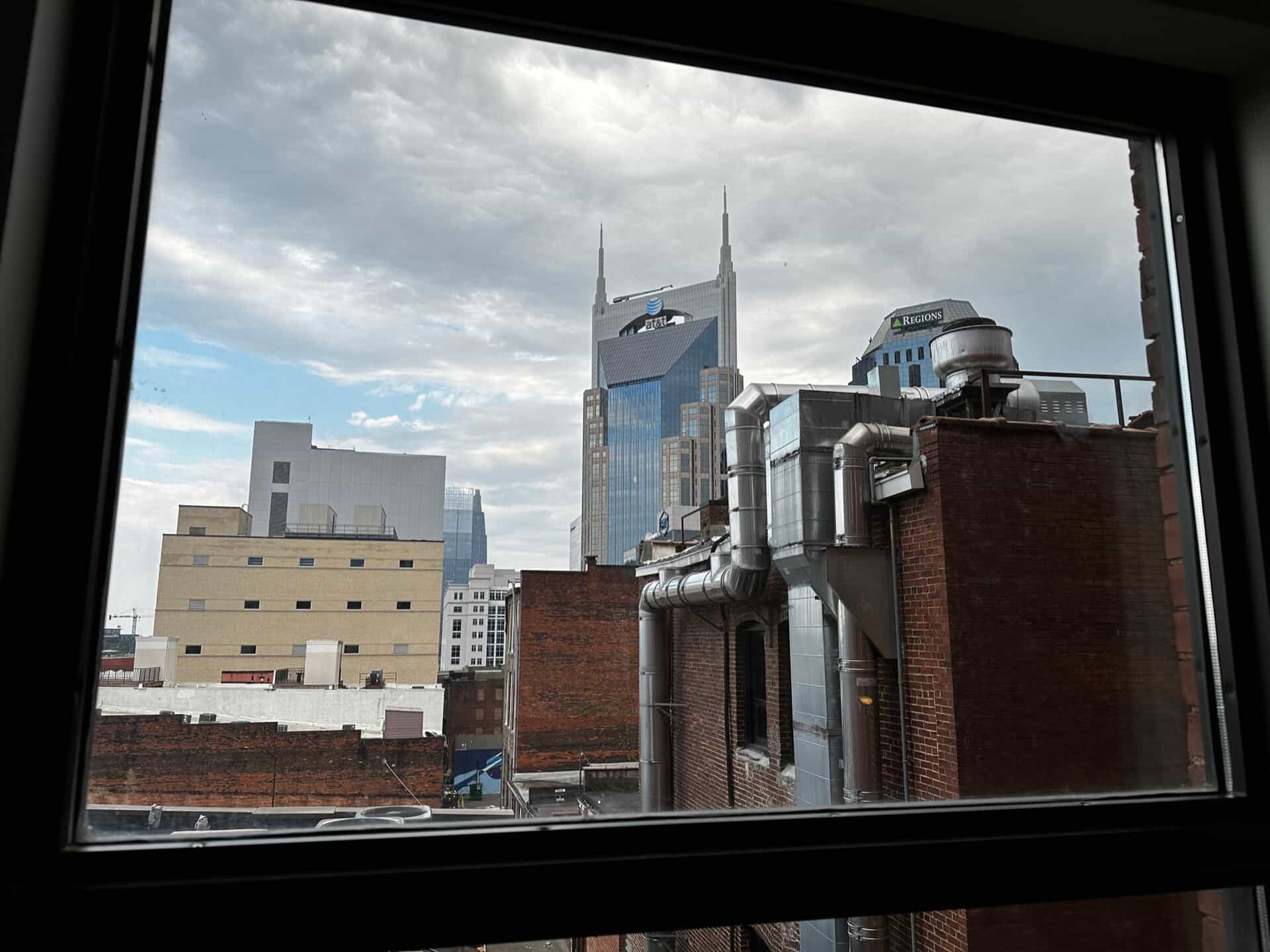 View of Nashville city buildings through a window, showcasing a prominent skyscraper with distinctive spires under a cloudy sky. Rooftops and ventilation systems create an intricate foreground.