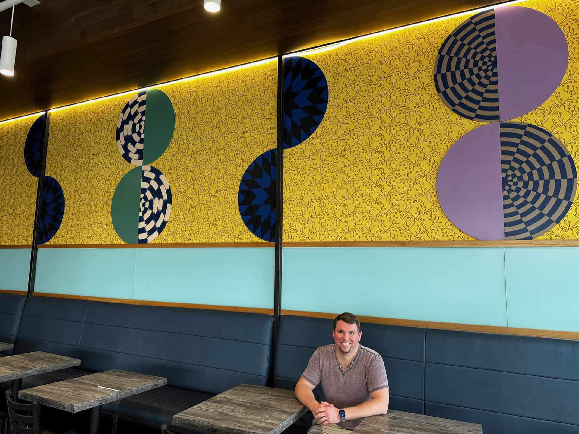 A man sits at a wooden table in a Nashville restaurant, surrounded by colorful geometric wall art with circles and patterns in blue, yellow, green, and purple.