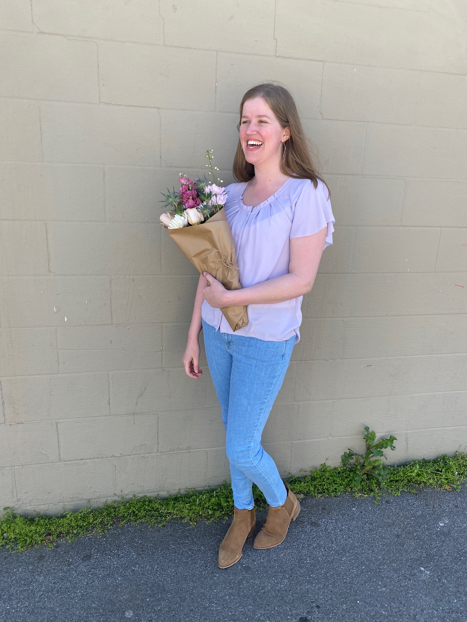 A woman in a lavender blouse and blue jeans stands against a beige wall in Nashville, holding a bouquet of flowers and smiling.