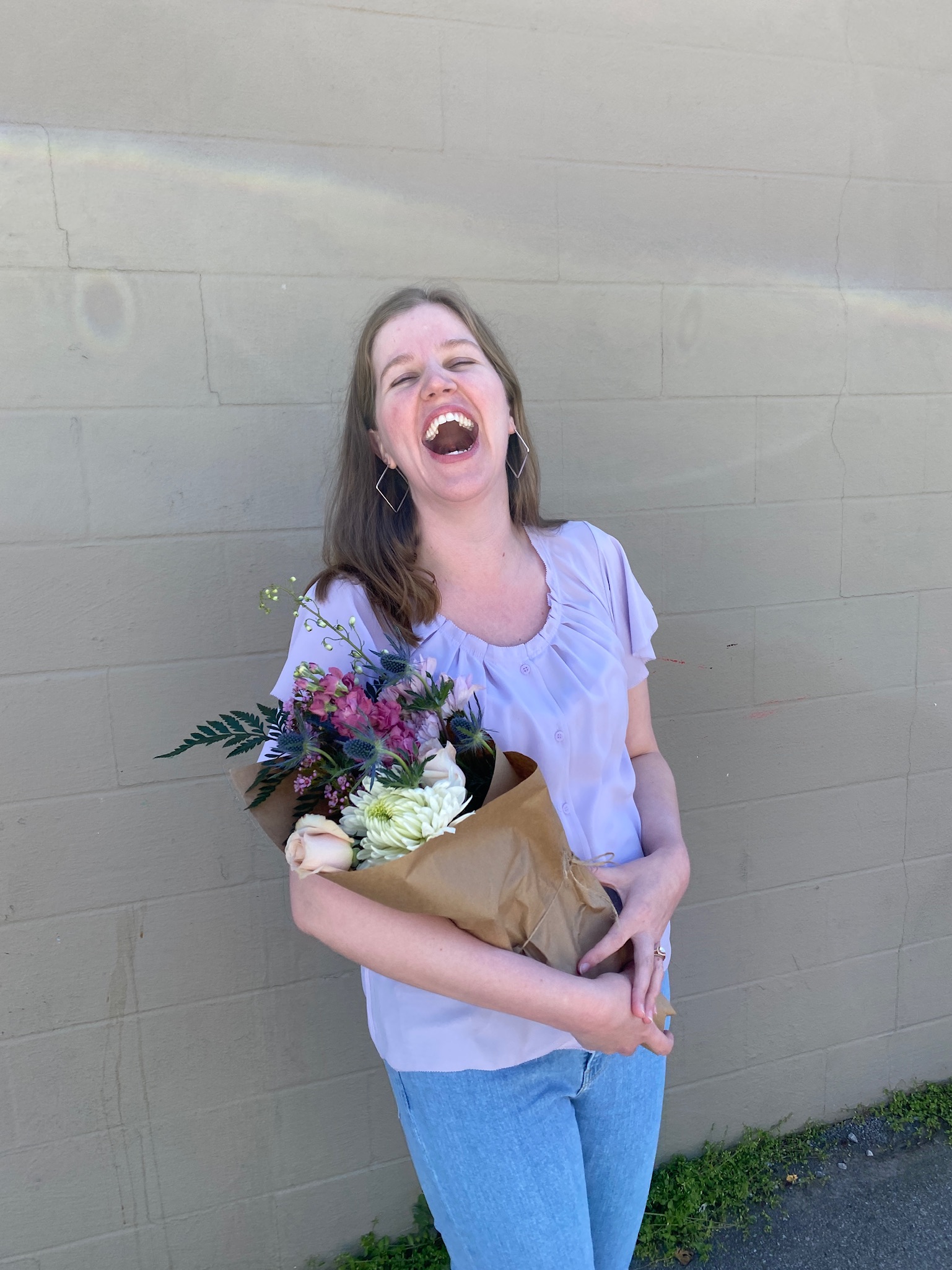 A woman in a light purple blouse and jeans stands against a beige wall in Nashville, holding a bouquet of flowers and laughing with her mouth open.