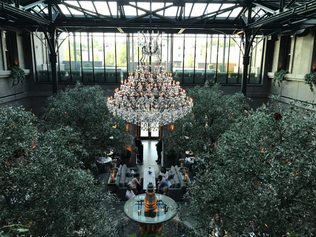 Spacious indoor restaurant with large windows, a central crystal chandelier, leafy green trees, and people dining at tables below.