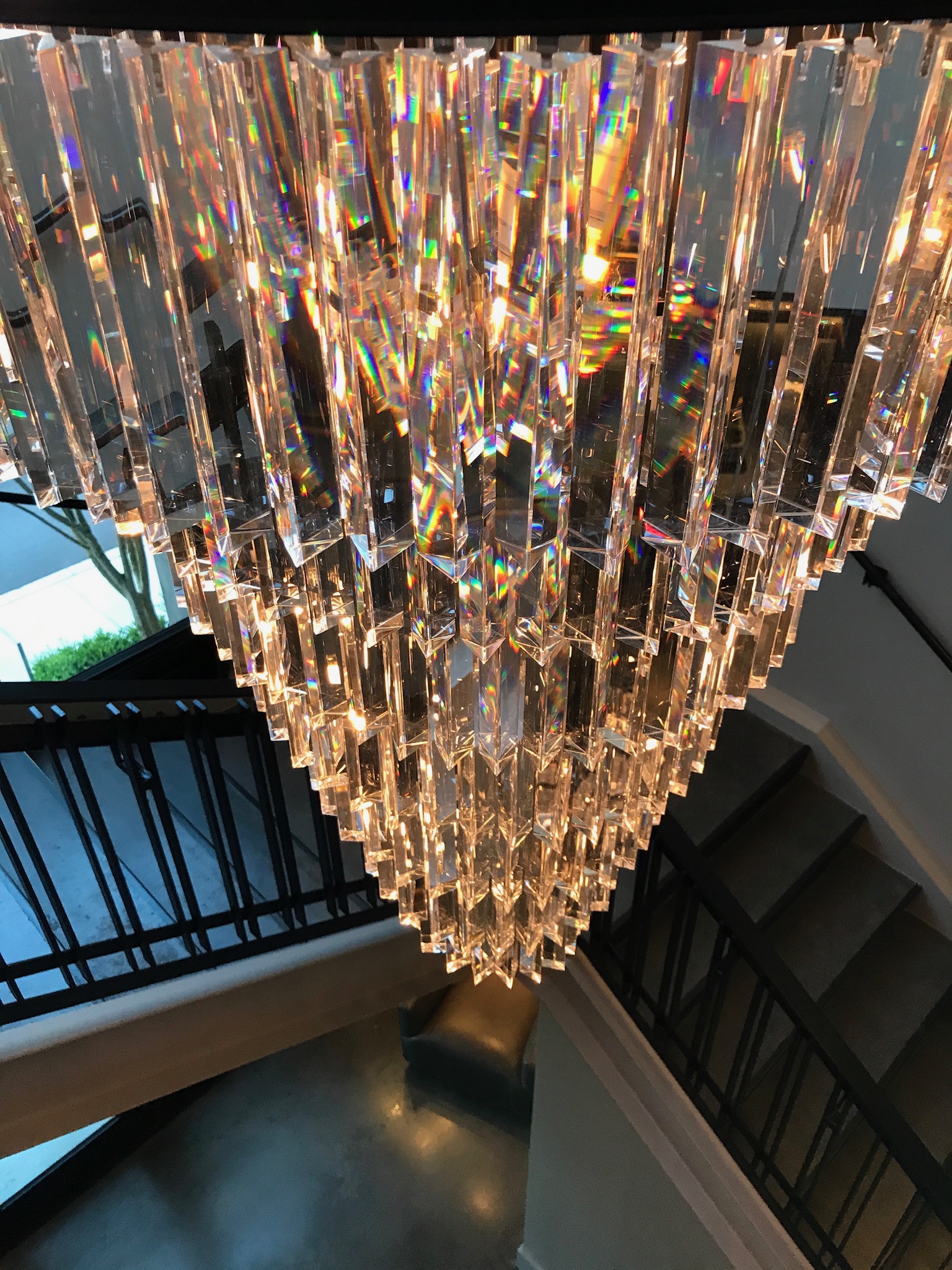 Large, triangular crystal chandelier hangs over a staircase, reflecting rainbow light patterns and illuminating the surrounding railings and steps.