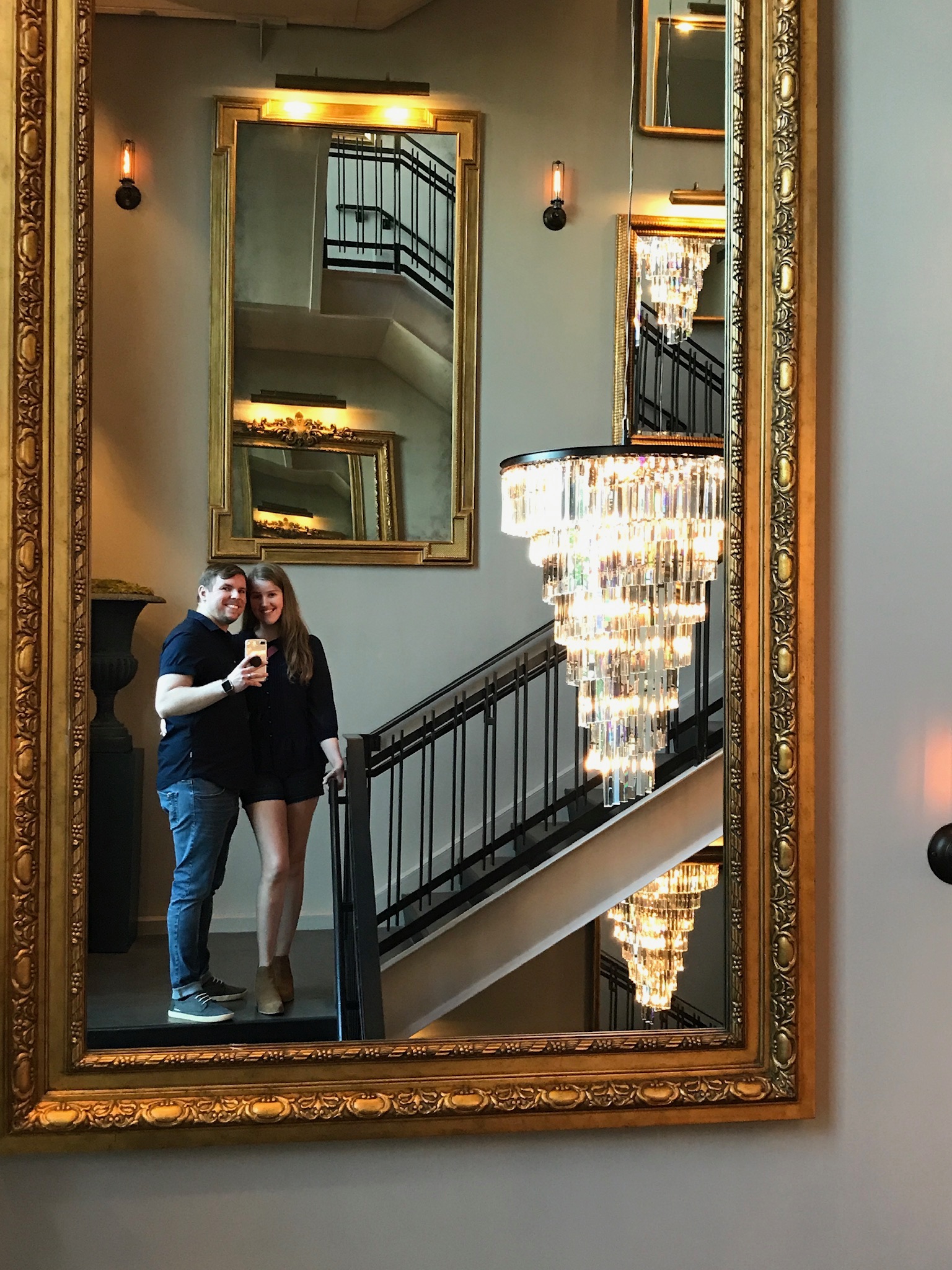 A couple stands on a staircase, posing for a selfie reflected in a large ornate mirror. A chandelier and multiple mirror reflections are visible in the background.