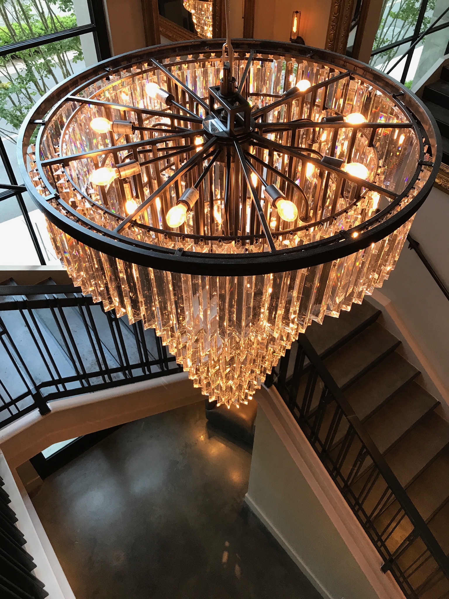 Large, illuminated crystal chandelier hanging above a stairwell with metal railings, reflecting light onto the surrounding walls and floor.