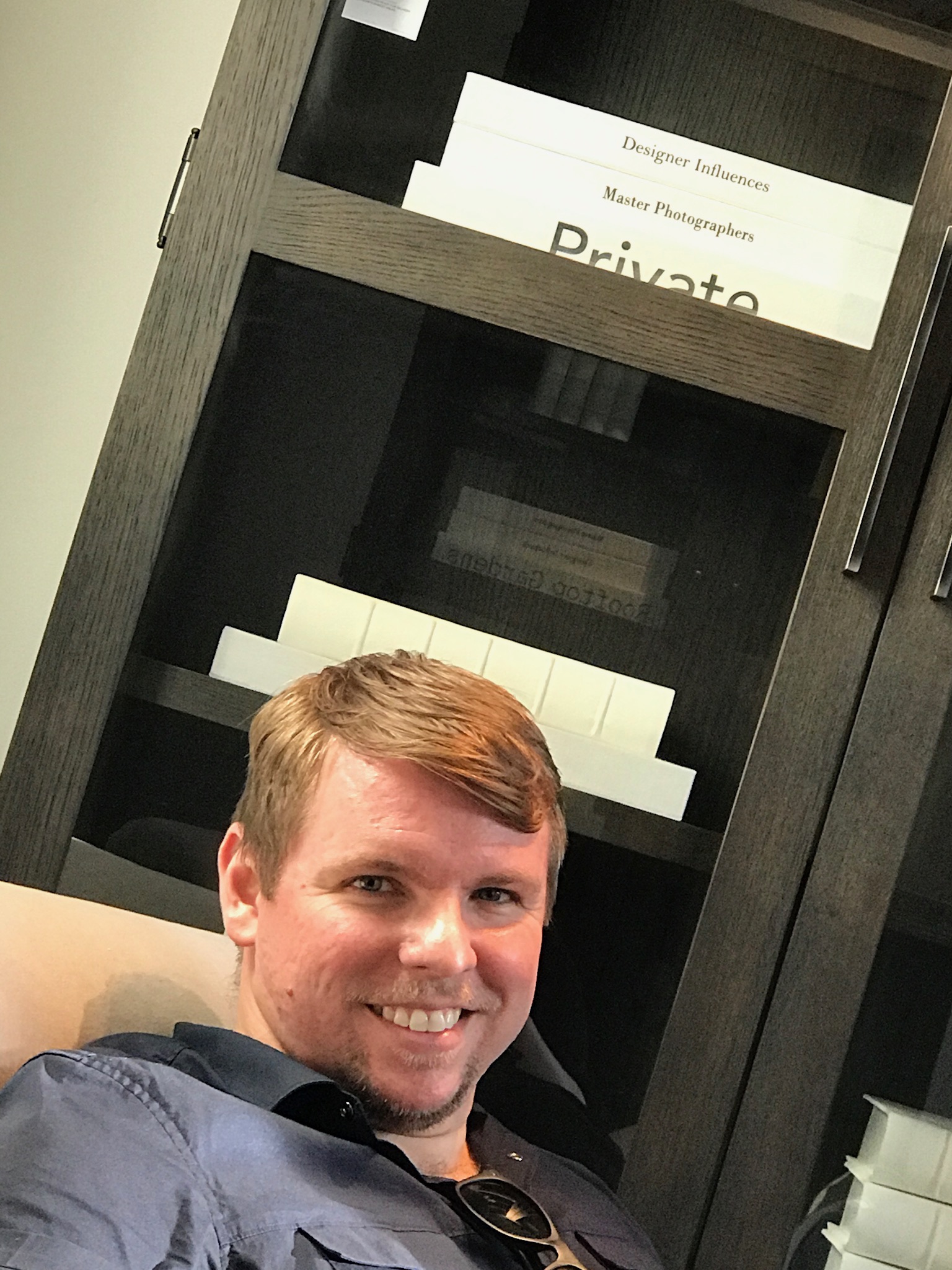 A man with light brown hair and a blue shirt smiles at the camera while sitting in front of a dark wooden cabinet with labeled shelves.