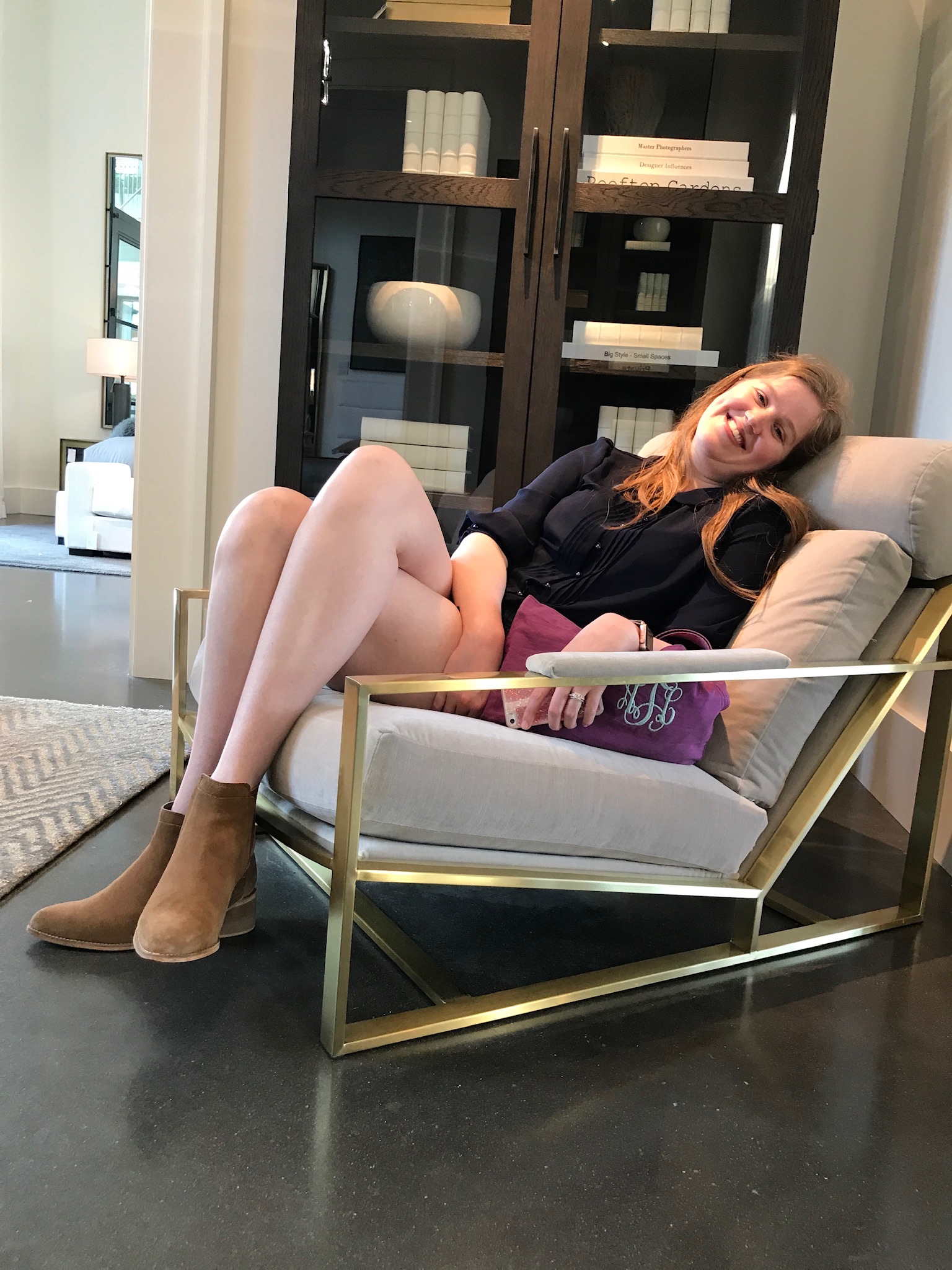 A woman sits relaxed in a modern armchair, smiling, holding a purple clutch, with bookshelves and a rug visible in the background.