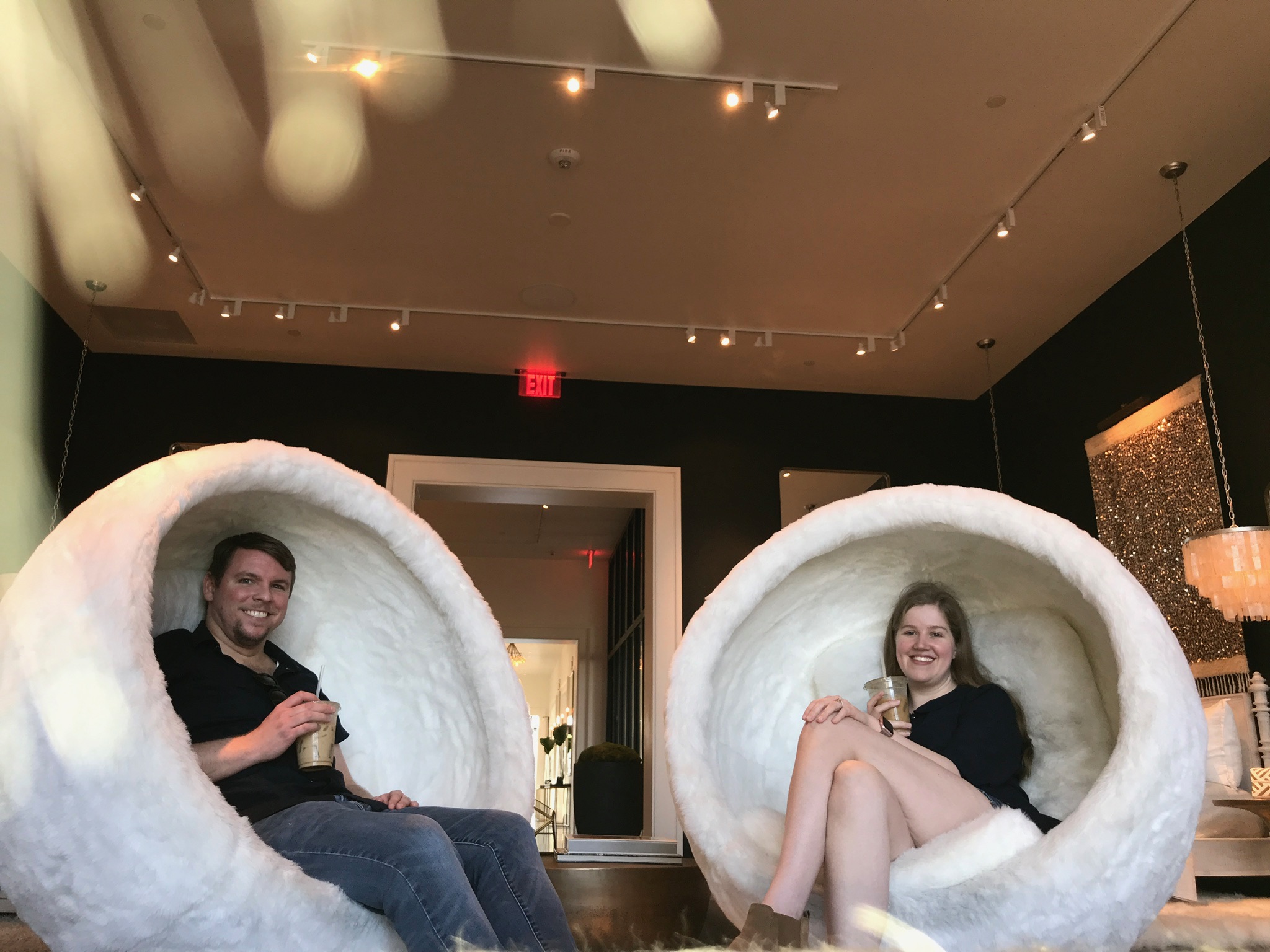 A man and a woman sit smiling in large, round, white, pod-shaped chairs indoors, each holding a drink.