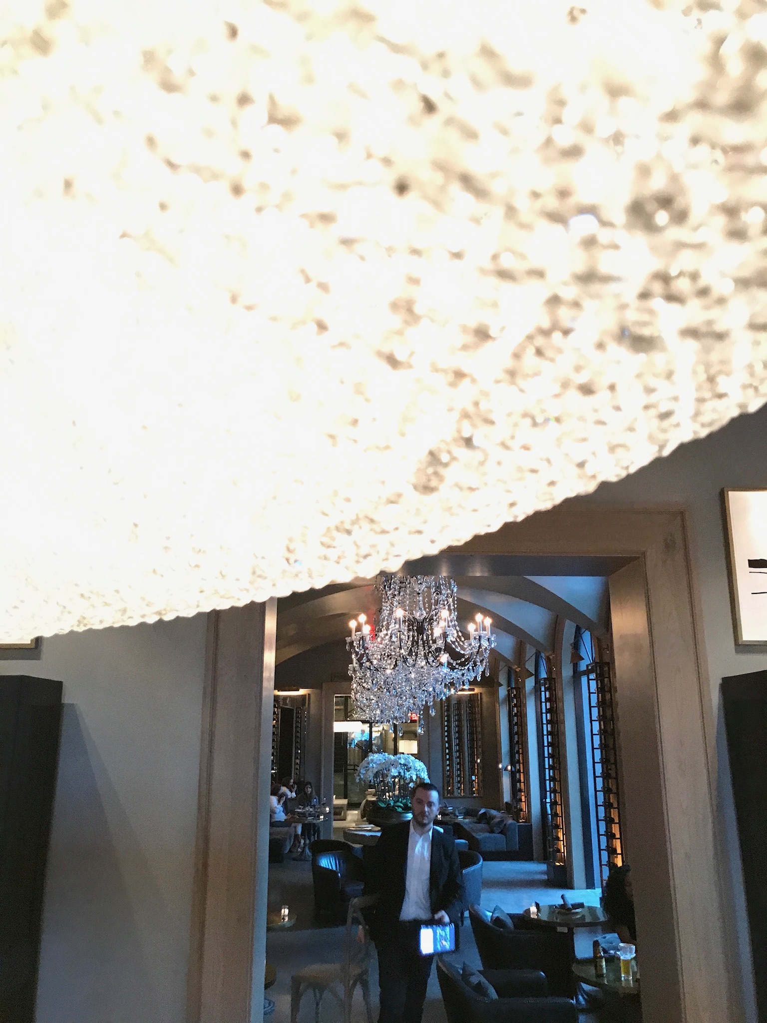 A man stands in a stylish lounge with a chandelier overhead, captured below a bright, textured ceiling feature that dominates the upper part of the image.