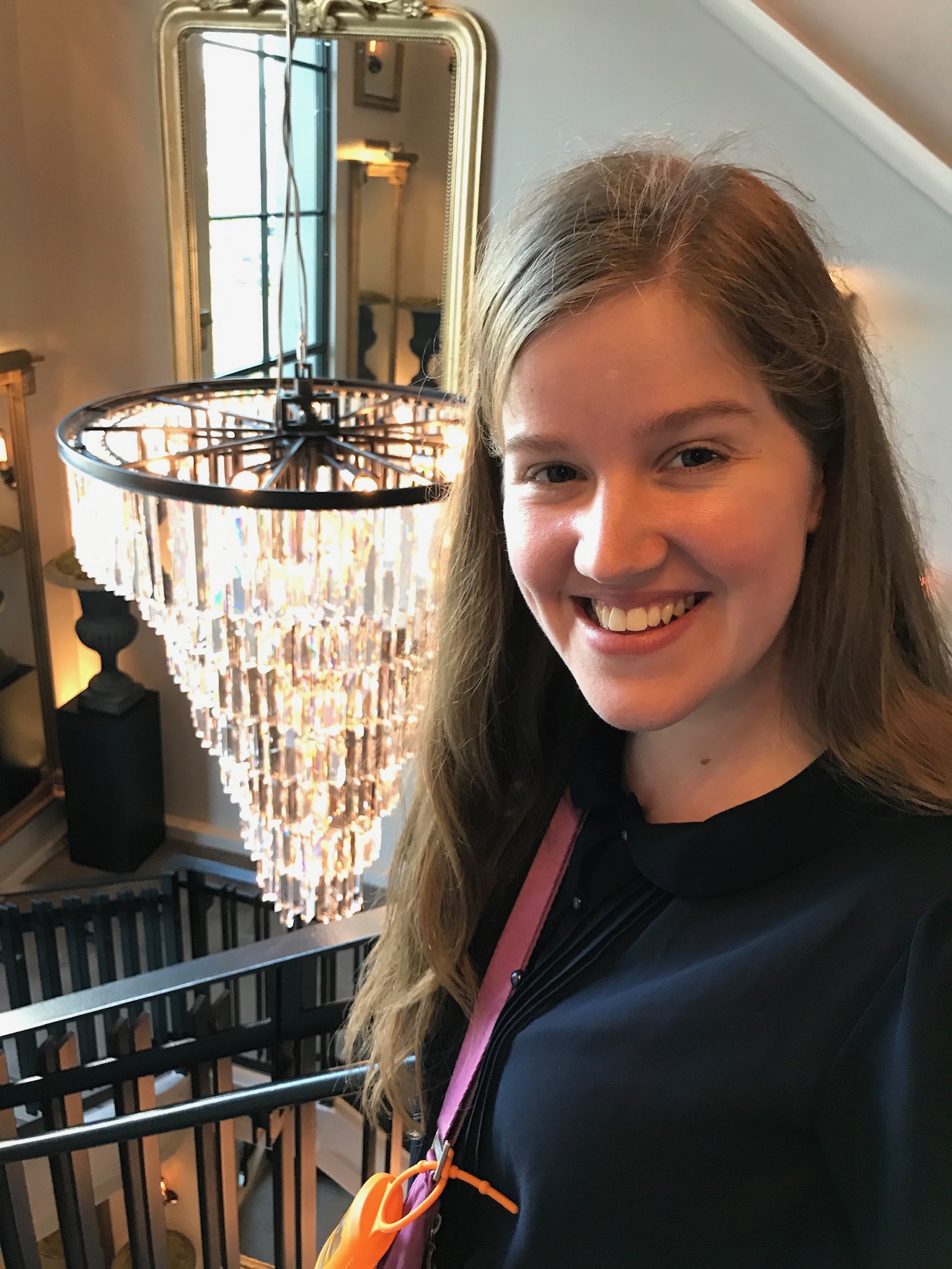 A young woman with long hair smiles at the camera, standing indoors by a staircase with a large, modern chandelier in the background.