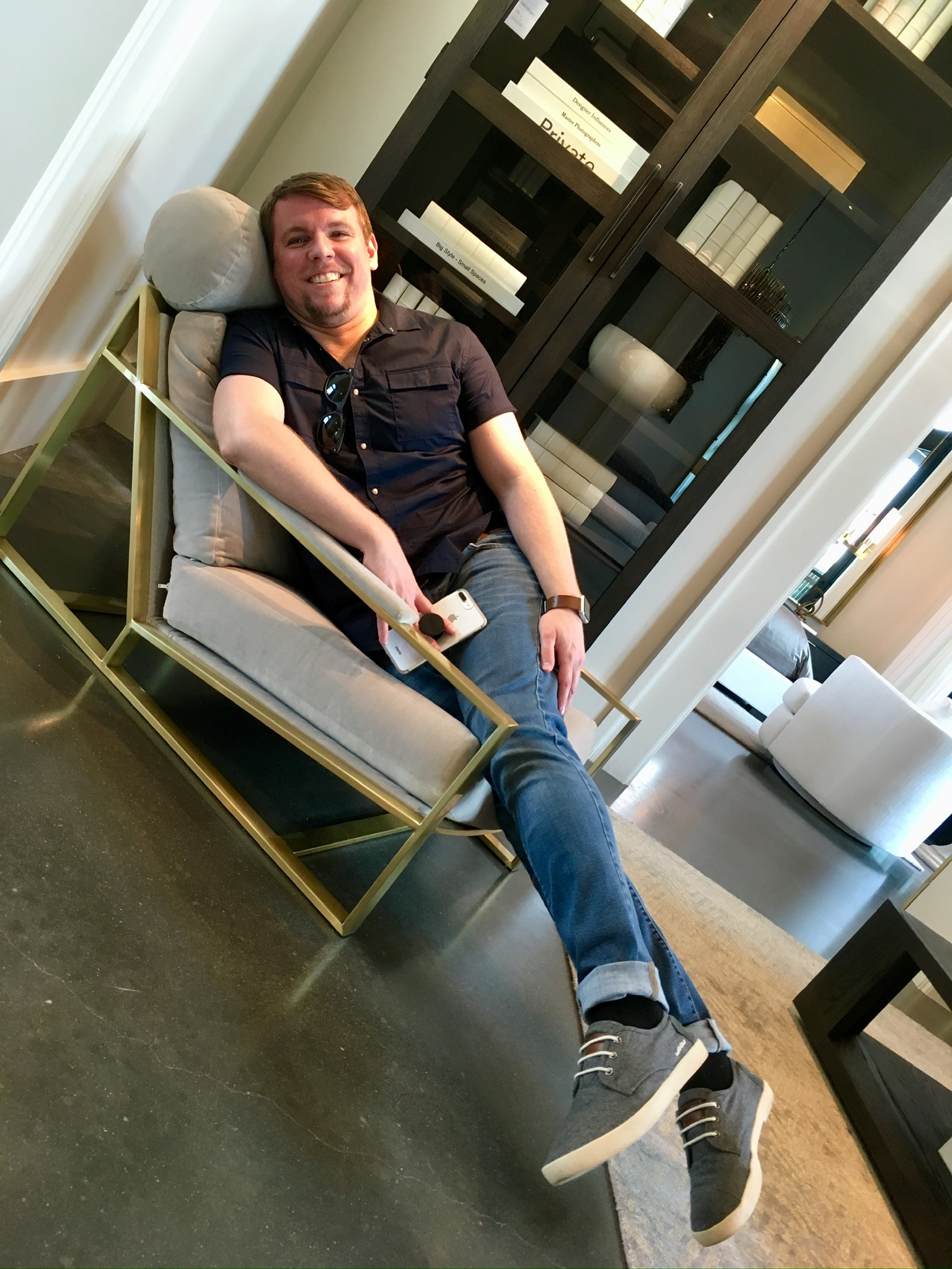 A man in casual clothes reclines in a modern armchair, holding a phone and sunglasses, in a contemporary living room showroom.
