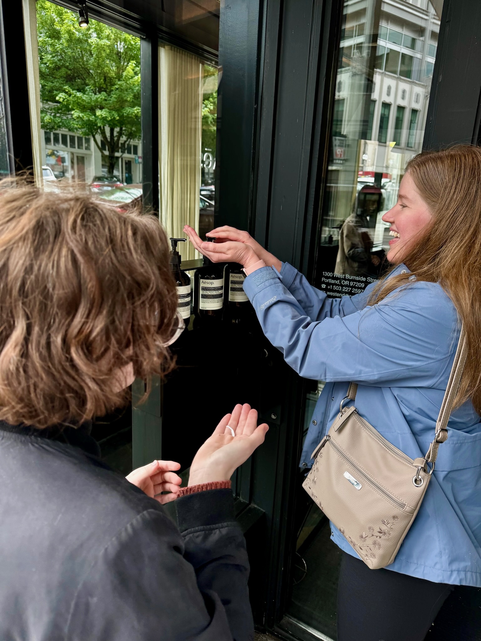 Two people stand near a doorway, one dispensing liquid from a bottle onto the other’s hands. Trees and buildings are visible outside through the glass.