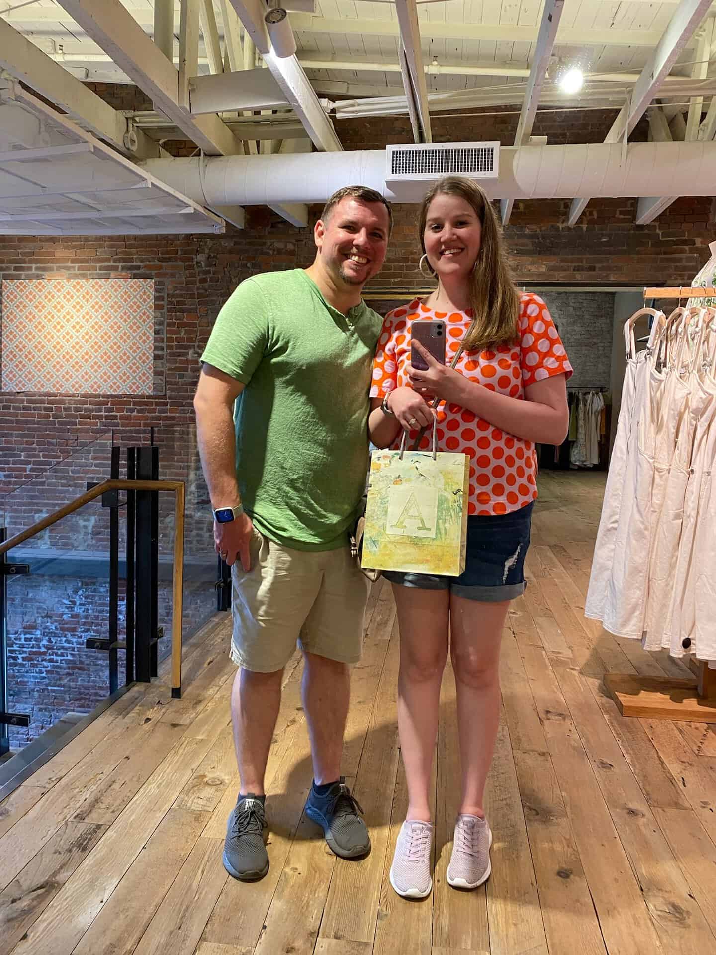 In a Nashville clothing store with wooden floors and brick walls, a man in a green shirt stands beside a woman in a red polka dot shirt holding a shopping bag.
