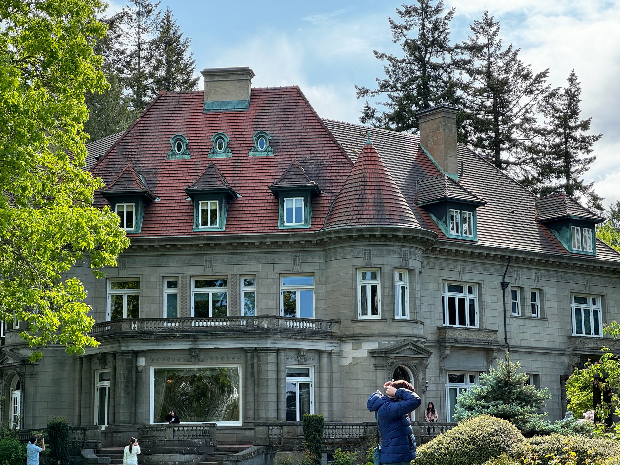 A large, historic mansion with red roof tiles, dormer windows, and ornate stonework, surrounded by trees and visitors in the foreground.