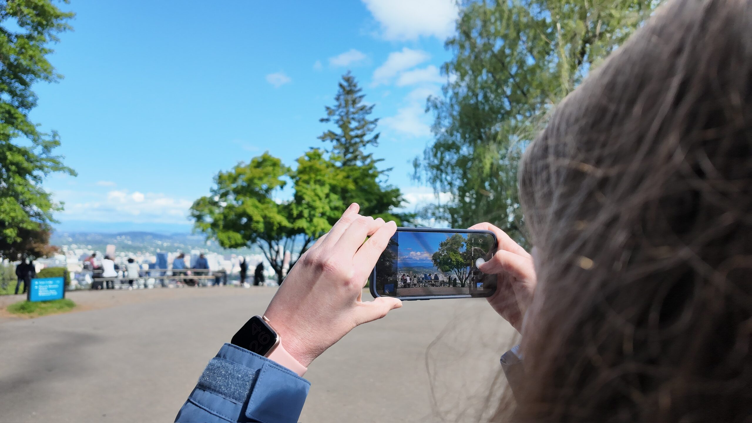 A person takes a photo of a scenic outdoor view with their smartphone, standing on a paved path surrounded by trees under a blue sky.