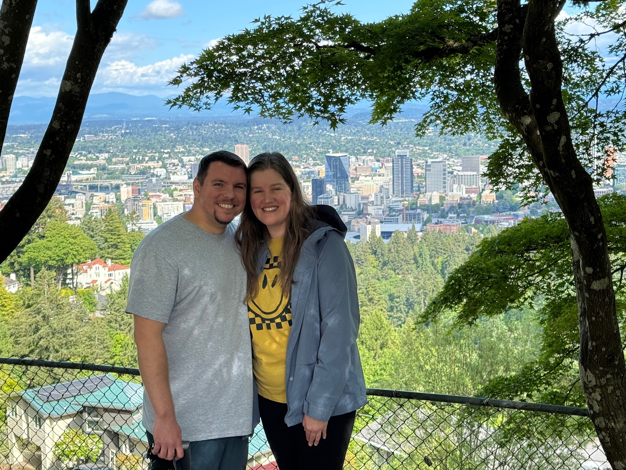 A man and woman stand smiling together in front of a fence with a view overlooking a cityscape, trees, and distant mountains.