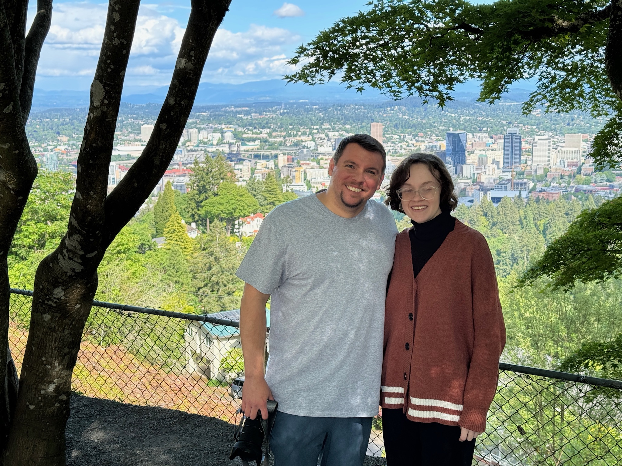 Two people stand smiling in front of a chain-link fence, with a cityscape, trees, and mountains visible in the background on a sunny day.