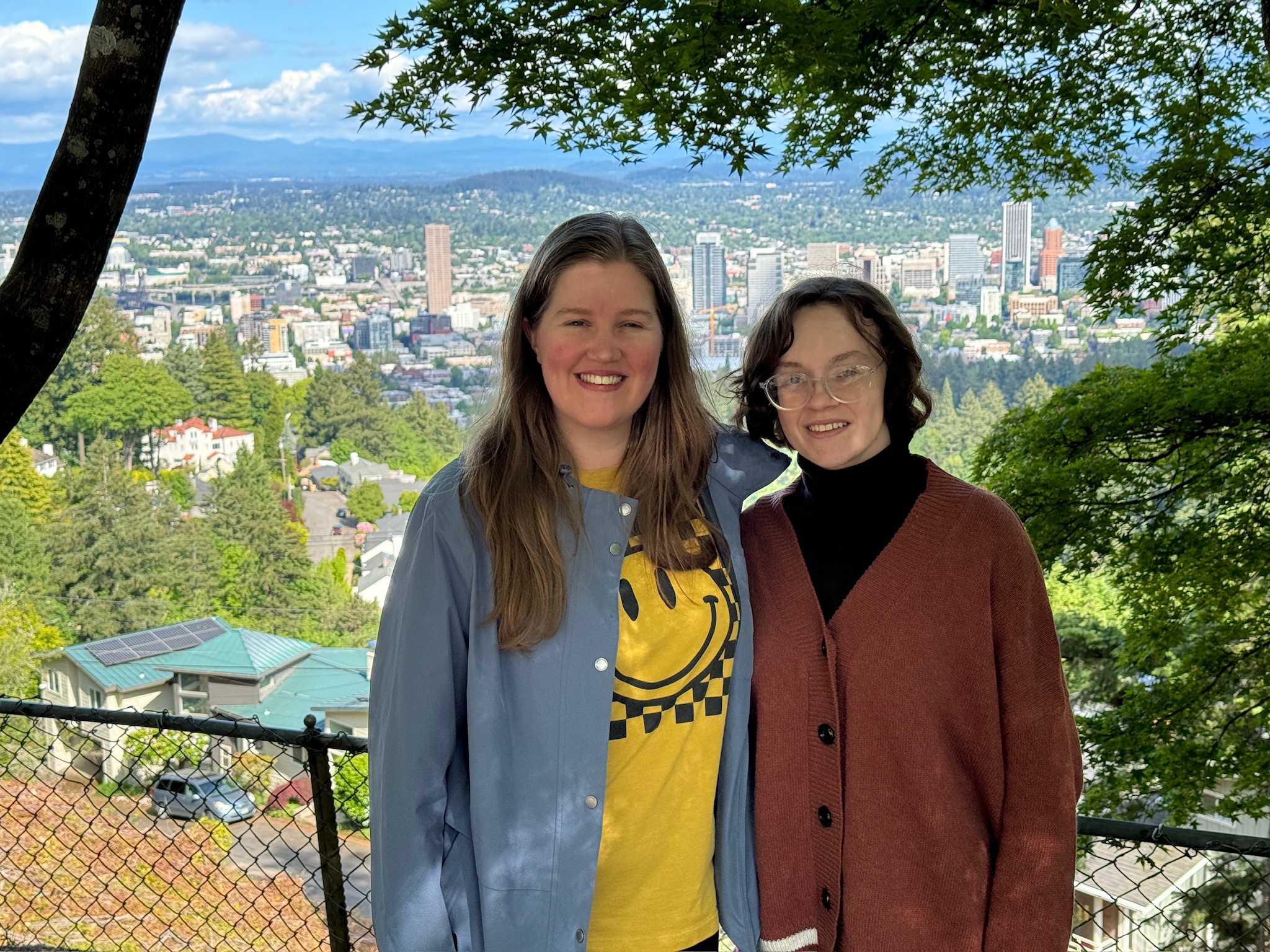 Two people stand side by side in front of a fence with a cityscape and greenery in the background, under a partly cloudy sky.