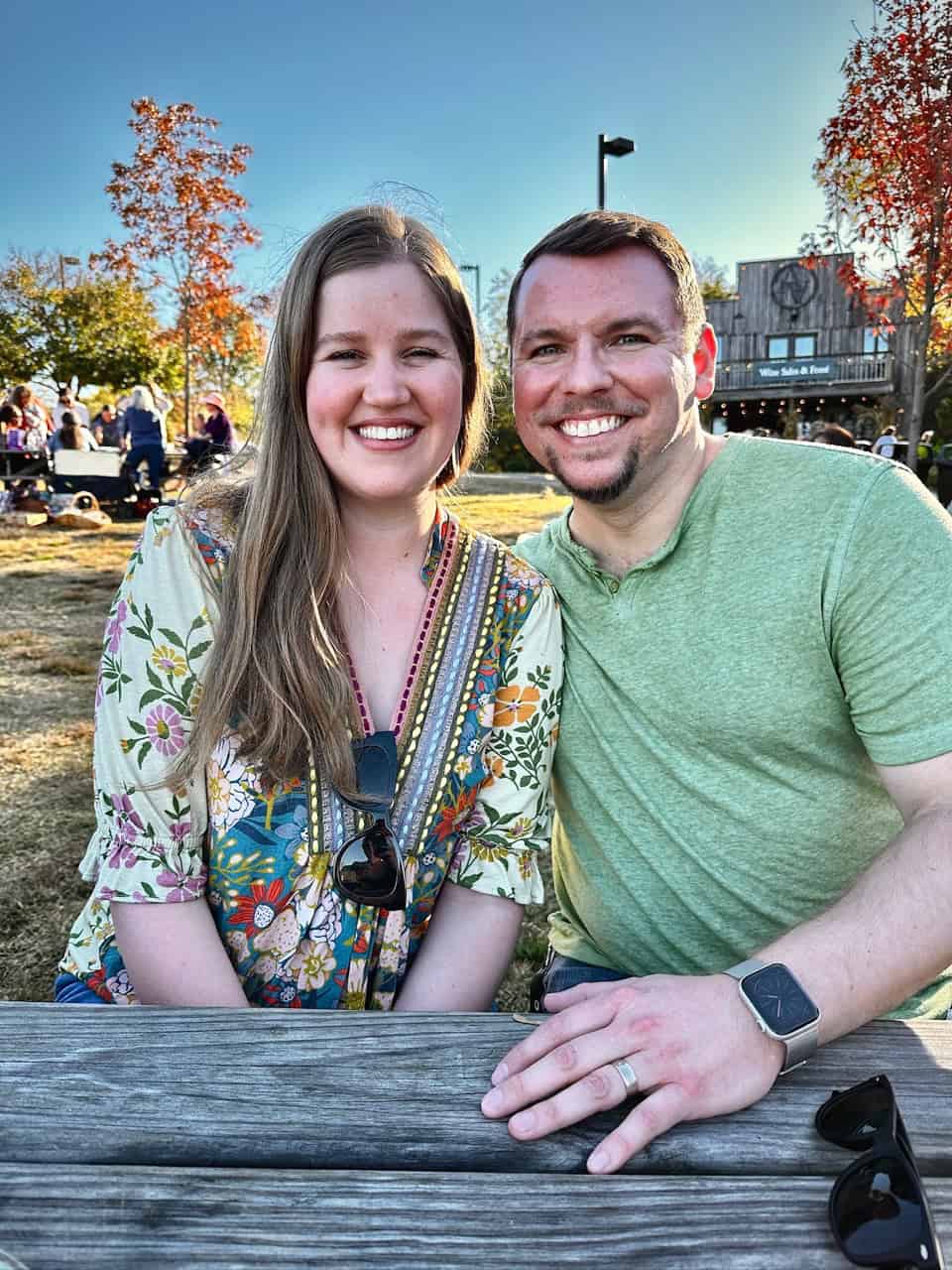 A man and woman sit at a picnic table in Nashville, smiling at the camera. The sky is clear, with trees adorned in vibrant autumn leaves painting a perfect background.