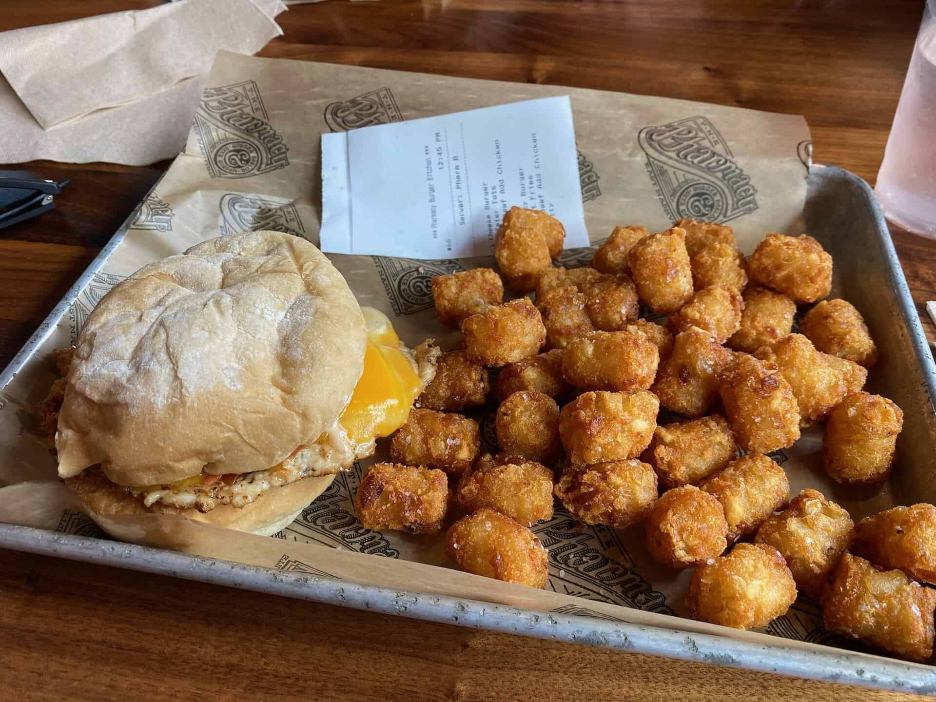 A sandwich with melted cheese is served on a metal tray alongside a pile of tater tots and a receipt on brown paper.