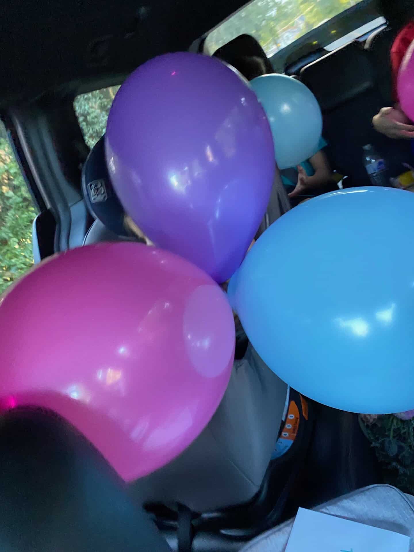A car interior with pink, purple, and blue balloons. Two children partially visible in the back seats.