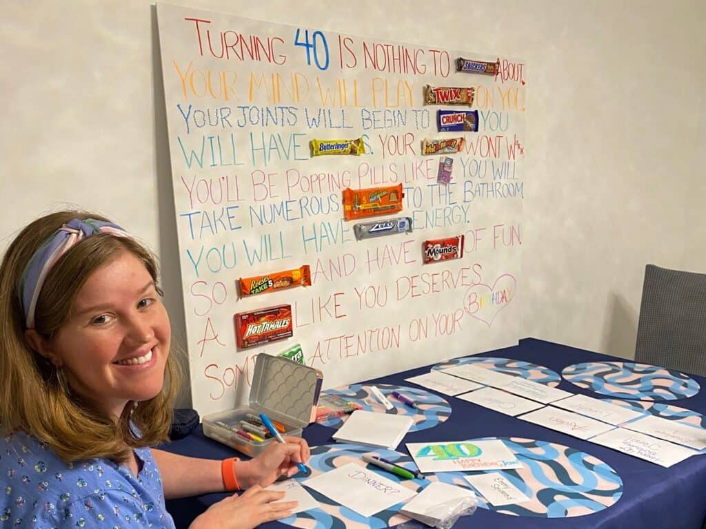 Woman seated at a table writing in cards, with a board in the background displaying candy bars and humorous text about Aunt Jenn's 40th birthday adventure.