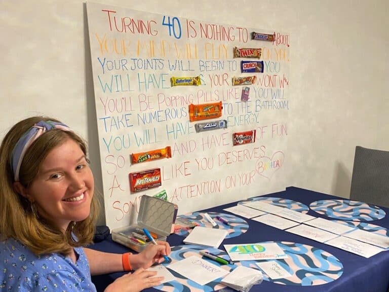 Woman seated at a table writing in cards, with a board in the background displaying candy bars and humorous text about Aunt Jenn's 40th birthday adventure.