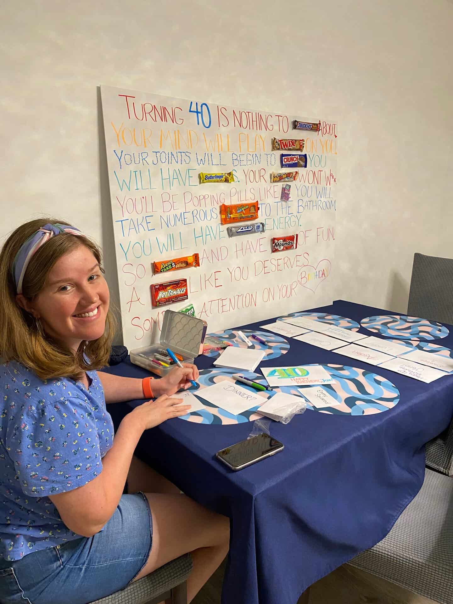 Smiling person sitting at a table with birthday cards and a poster. The poster humorously mentions turning 40 and includes candy bars as part of its design.