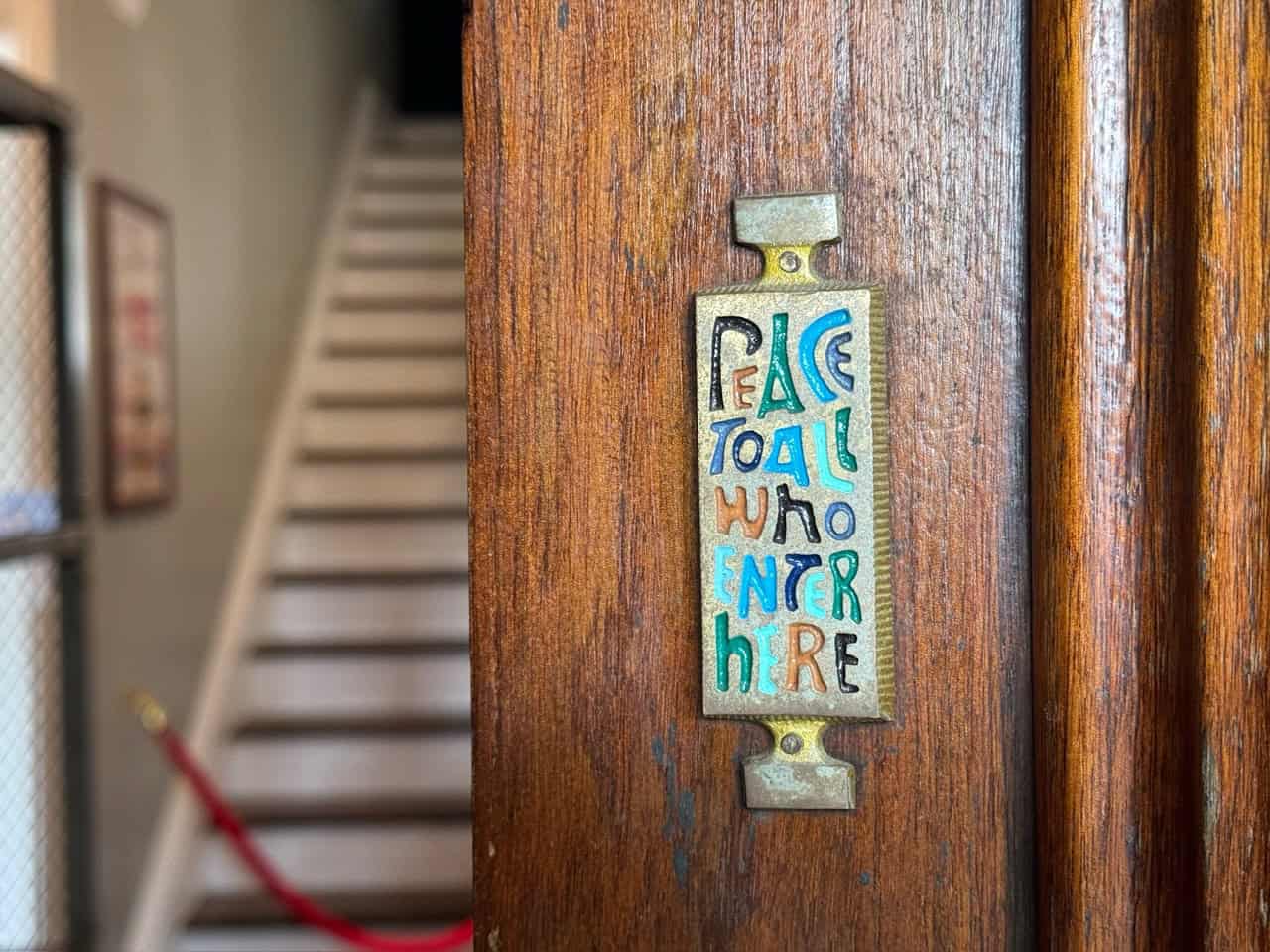Plaque on a wooden door reads "Peace To All Who Enter Here" in colorful text, greeting you with Southern charm. In the background, stairs and a red rope barrier hint at Nashville's warm hospitality waiting beyond.