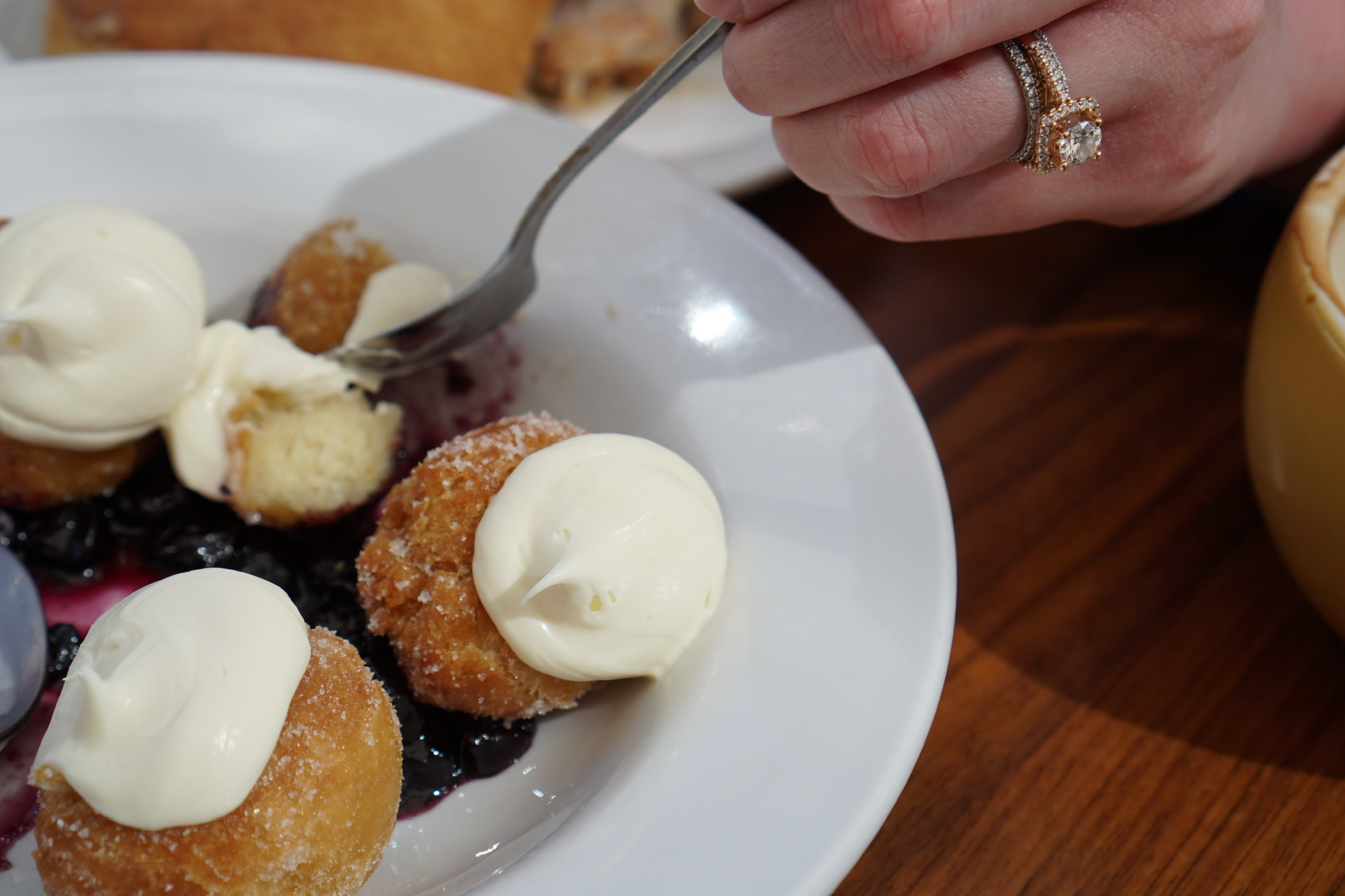 A hand with rings holds a fork over a plate of sugar-coated doughnuts topped with cream and blueberries, next to a cup on a wooden table.