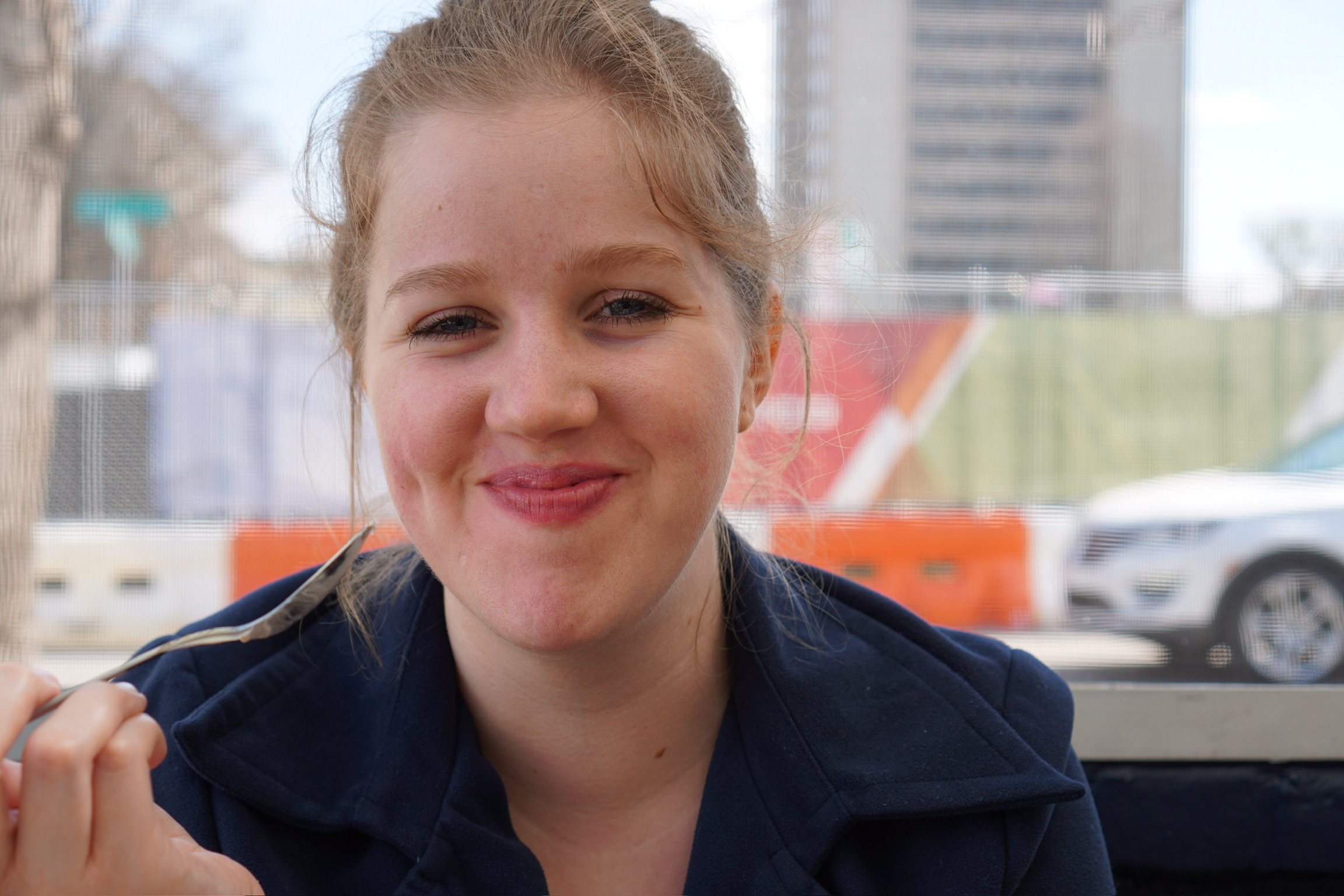A young woman with light hair smiles while holding a fork, sitting outdoors with construction barriers and cars in the background.