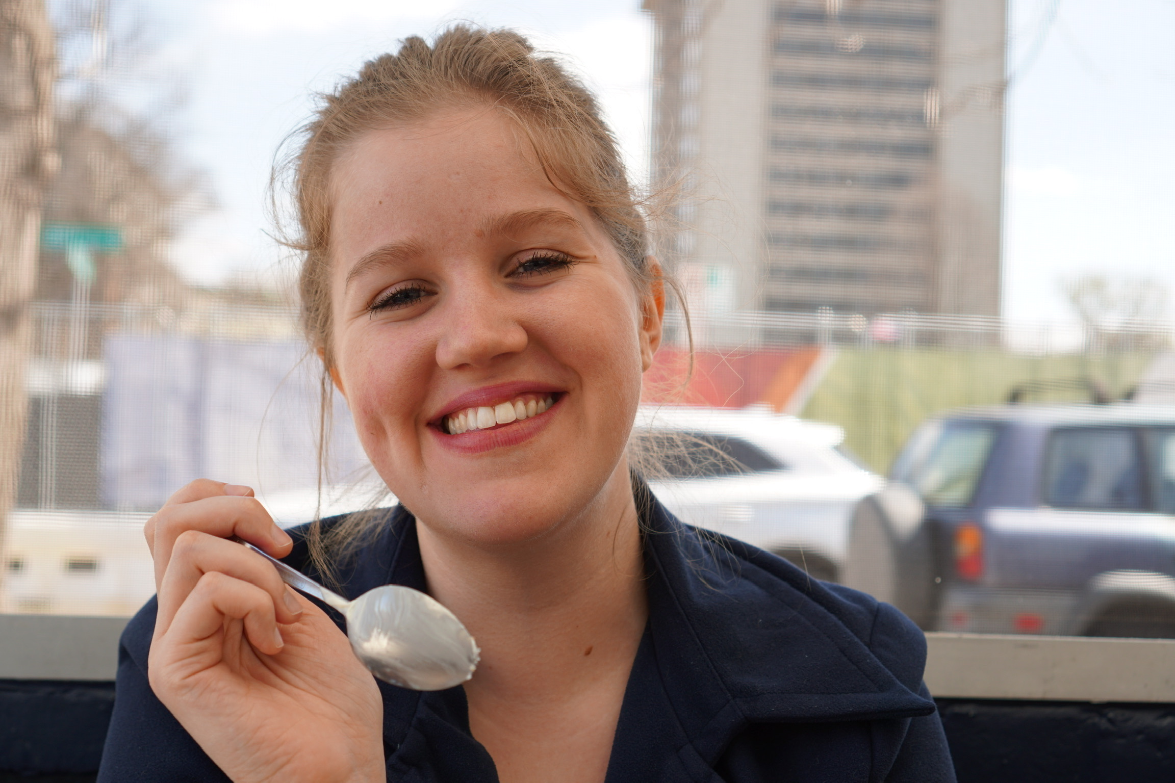 A woman smiling at the camera holds a spoon with a white substance, sitting outdoors with buildings and parked cars in the background.