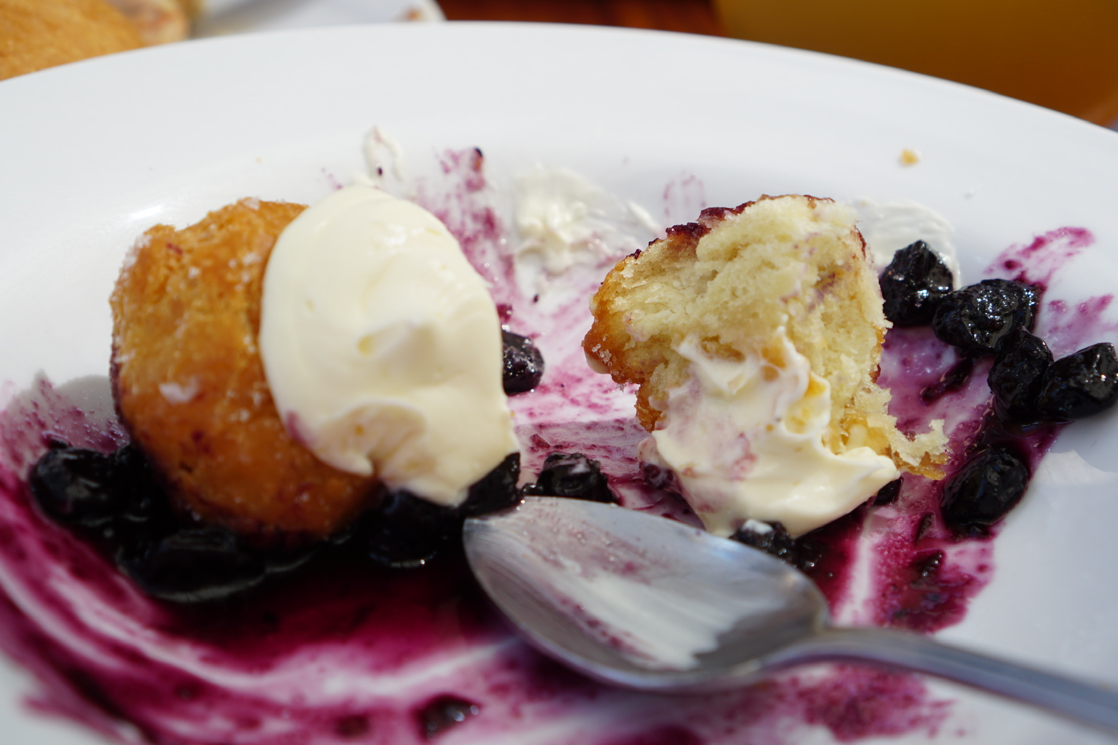 A partially eaten dessert with cream, fried dough, blueberries, and sauce on a white plate with a spoon.