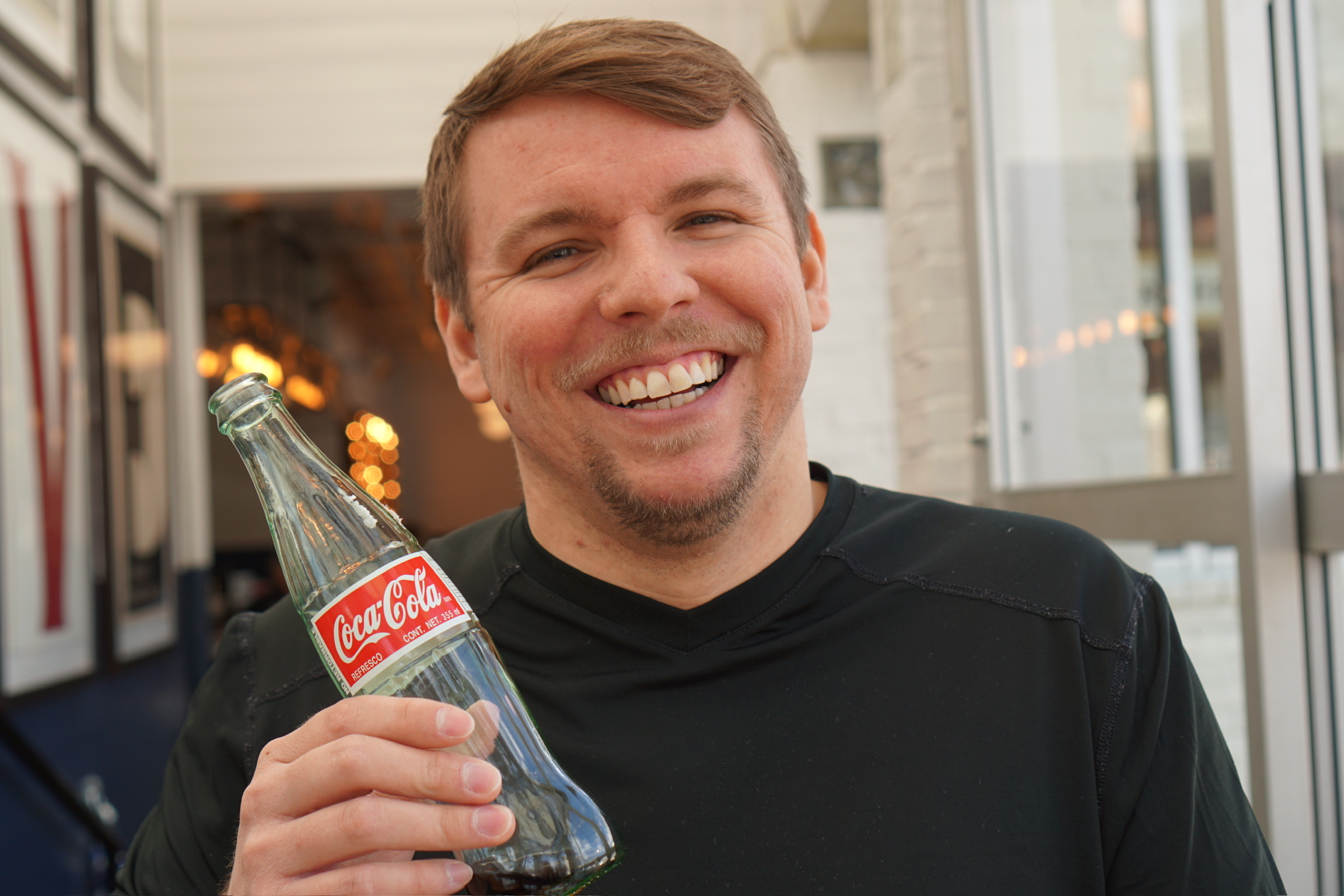 Man smiling and holding a glass bottle of Coca-Cola, standing indoors near a window.
