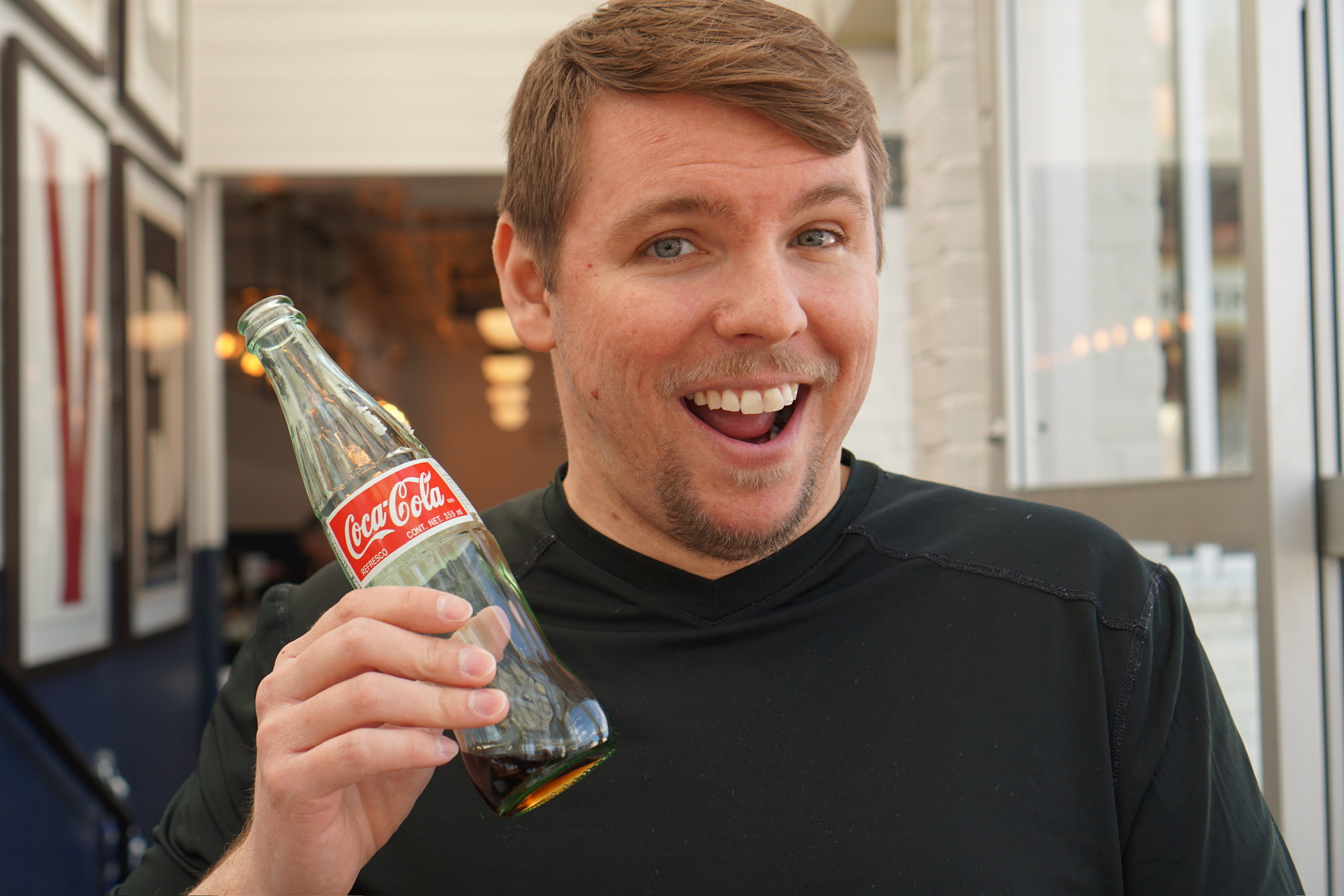 Man in a black shirt smiling and holding a glass bottle of Coca-Cola in front of a modern, well-lit indoor setting.